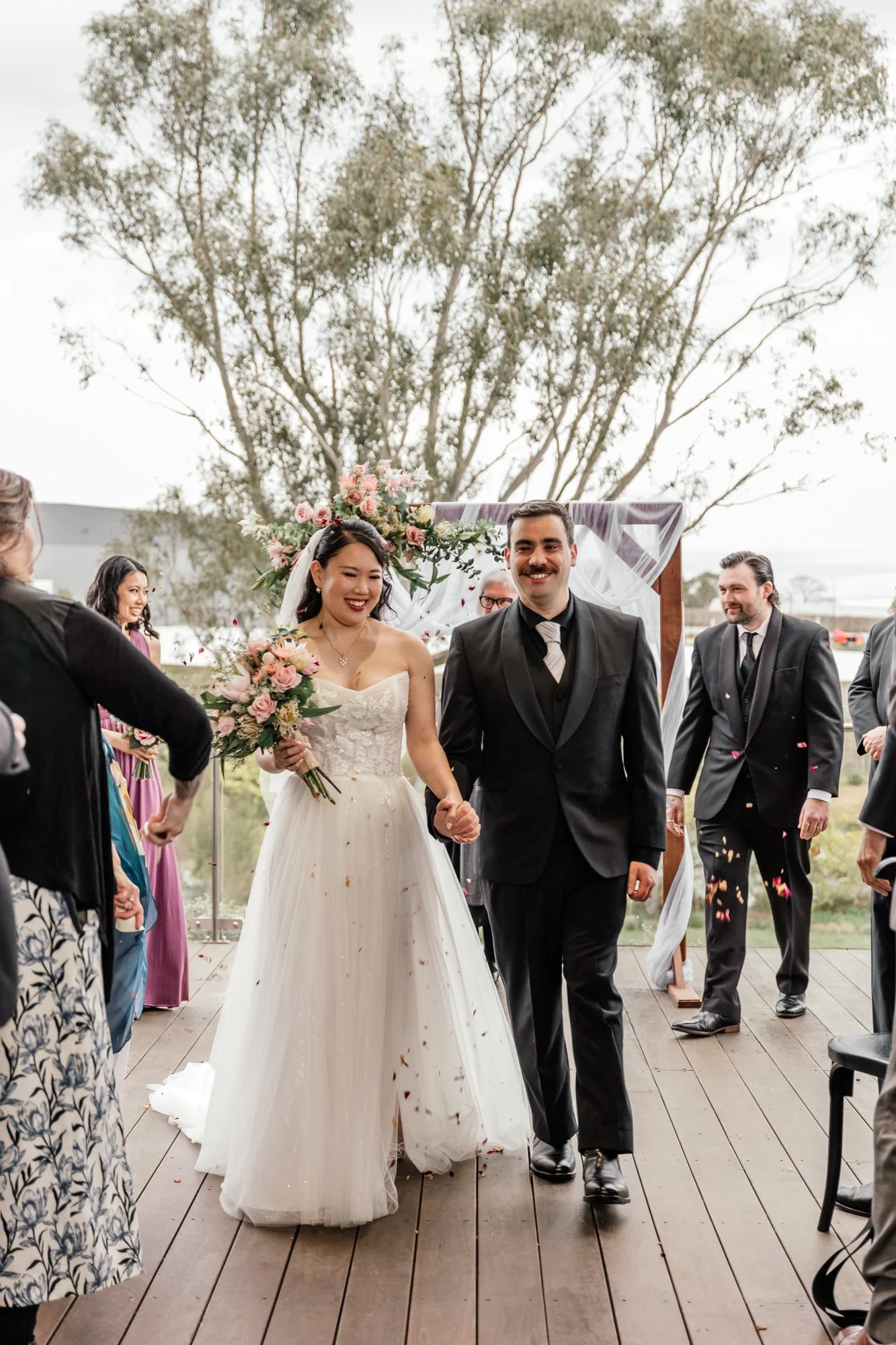 A newly married couple walking hand in hand down a wooden aisle outdoors, surrounded by friends and family, with a decorated arch and trees in the background.
