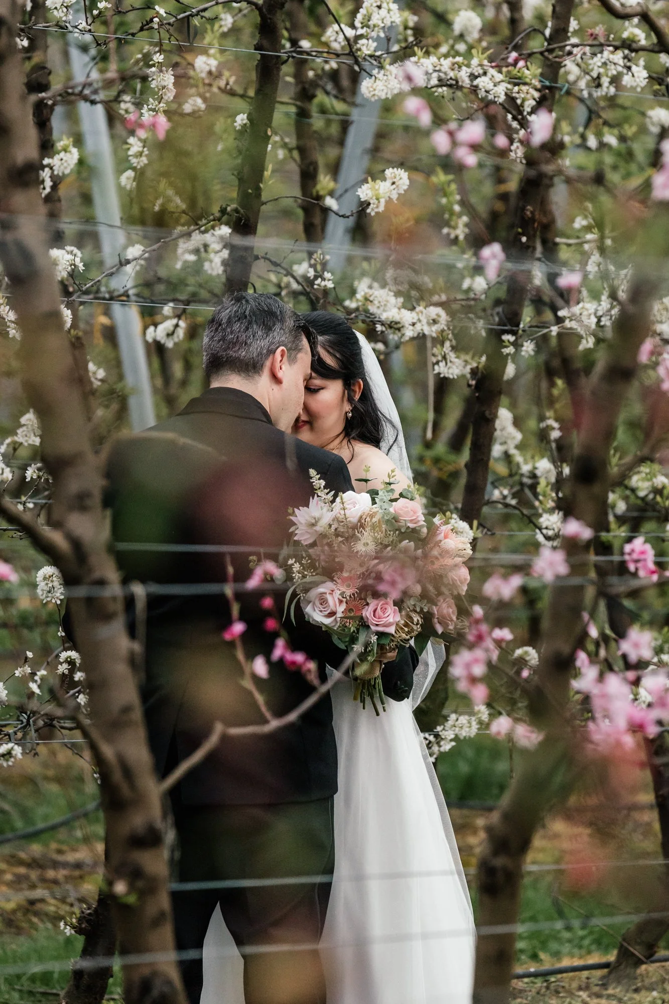 A newlywed couple sharing an intimate moment in a blooming orchard, surrounded by pink and white blossoms, with the bride holding a bouquet of roses and other flowers.