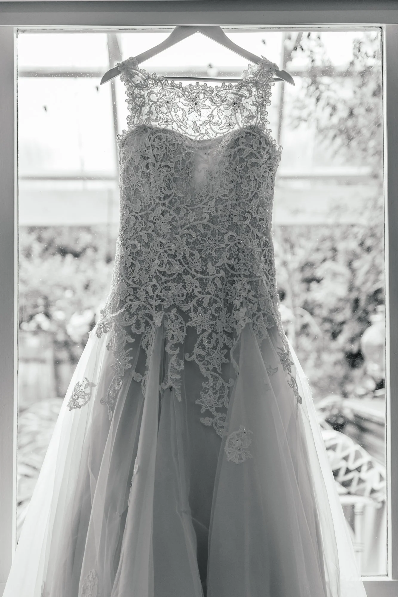 A wedding dress with intricate lace details hangs on a hanger in front of a window, with sunlight shining through.