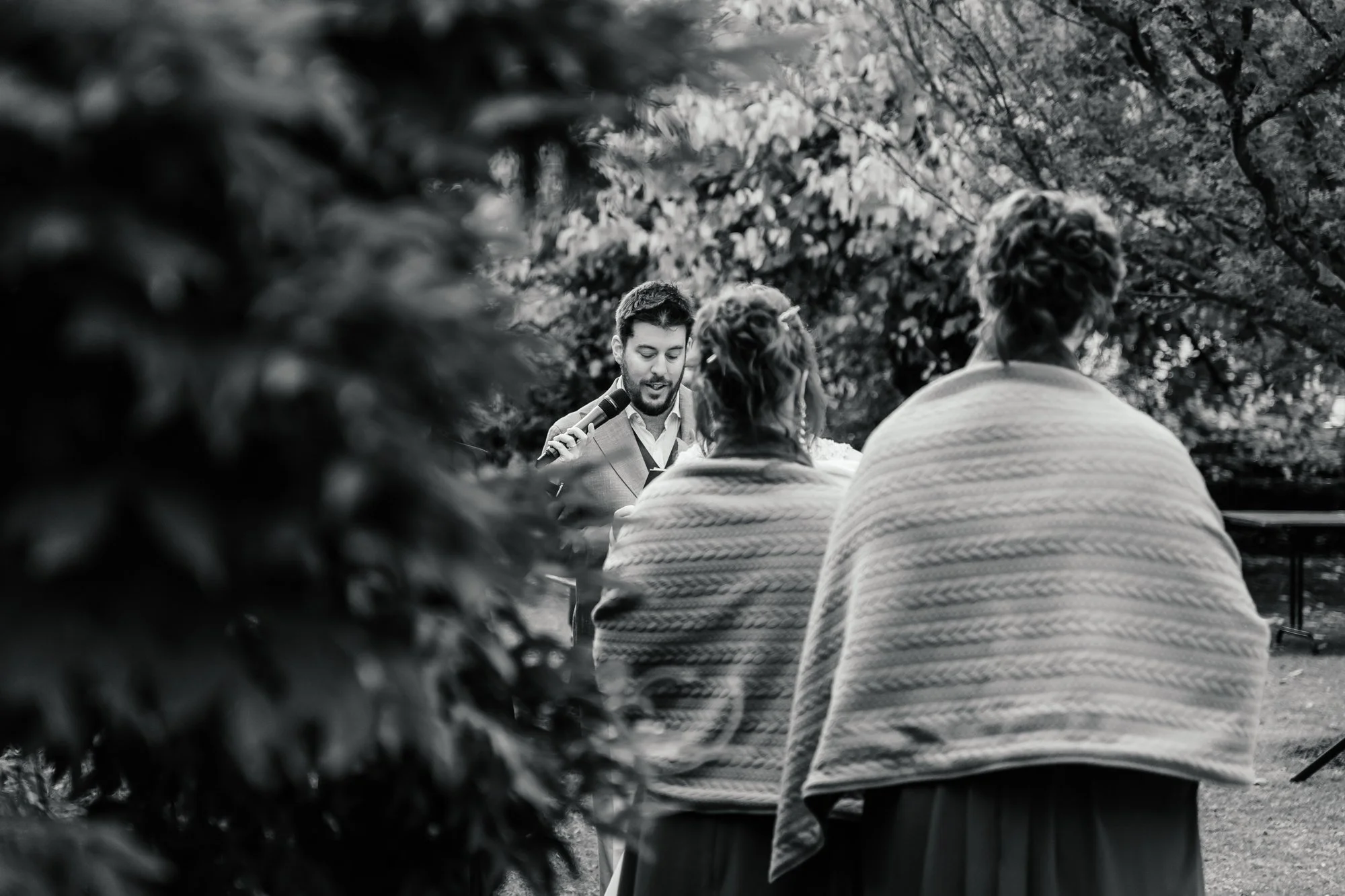 A man in a light-colored suit is speaking into a microphone during an outdoor event, with two women sitting in front of him, wrapped in blankets. The scene is surrounded by trees and foliage.