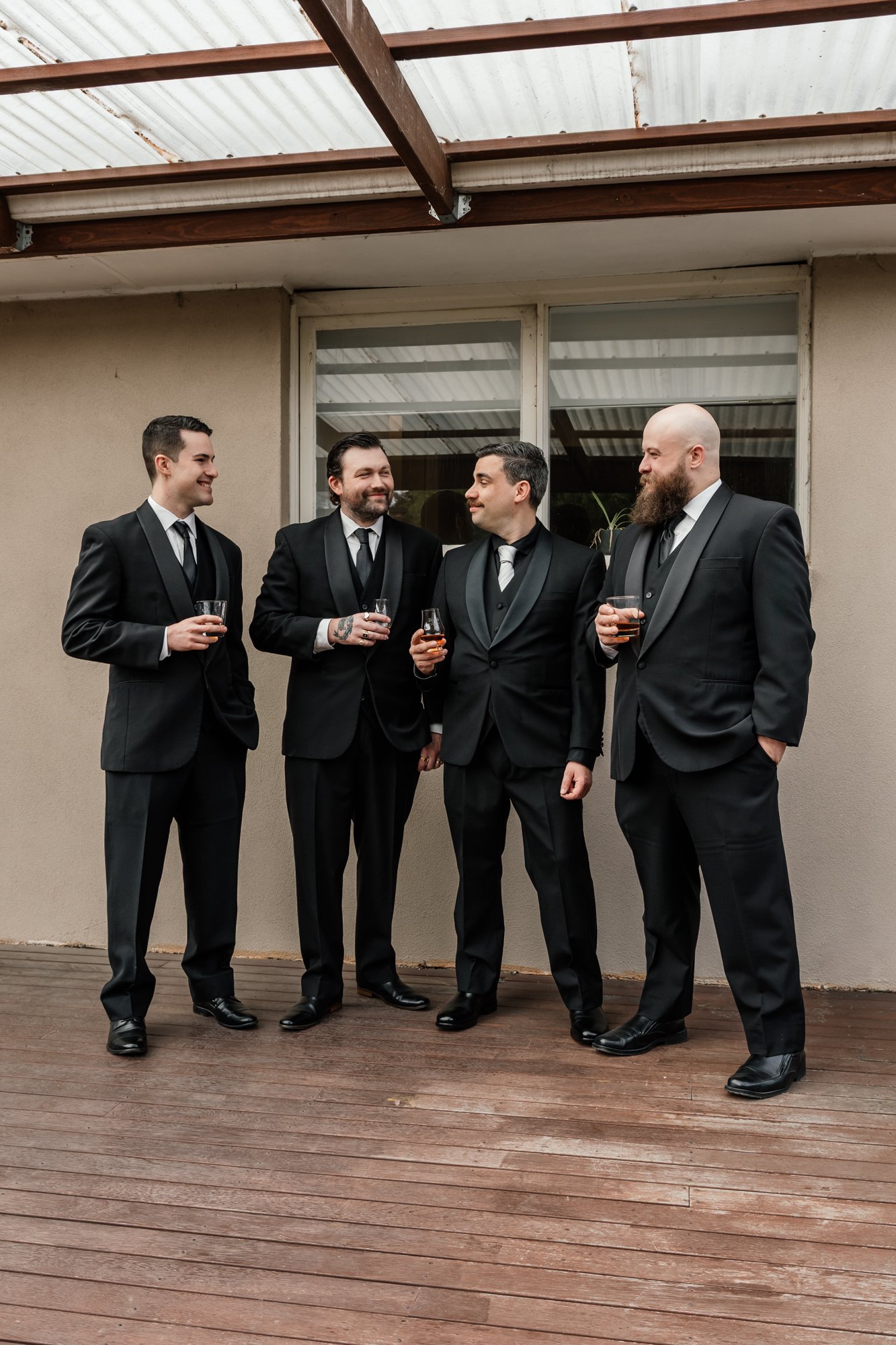 Four men in black tuxedos with ties, holding drinks and conversing on a wooden deck outdoors near a building with large windows.