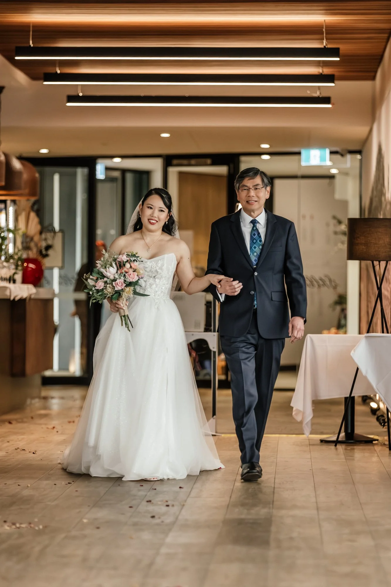 A bride in a white wedding dress holding a bouquet of pink and white flowers, walking arm-in-arm with a man in a dark suit and glasses, in a decorated indoor venue.