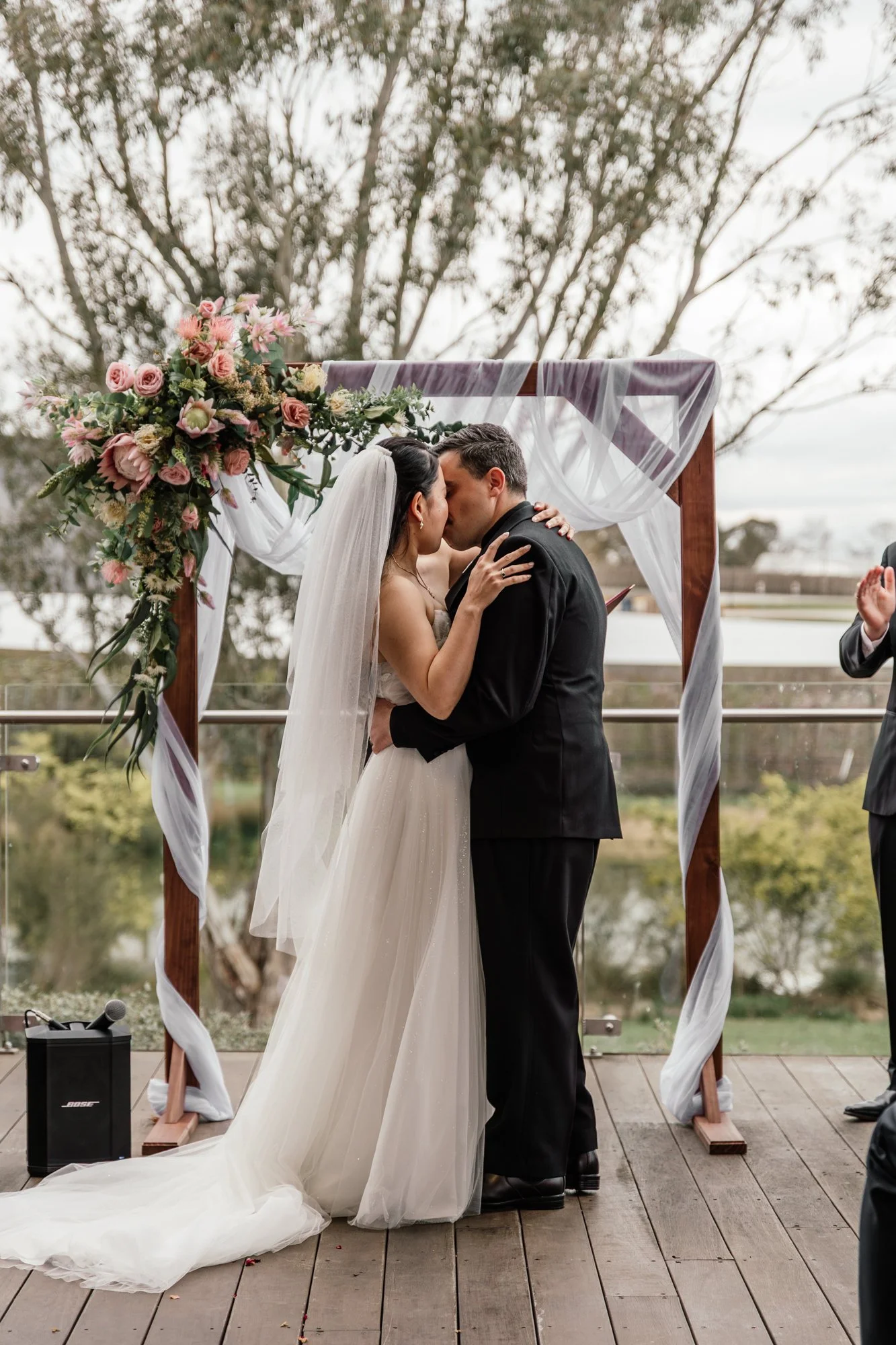 A bride and groom kiss during their outdoor wedding ceremony under a decorated arch with pink and white flowers, sheer white fabric, and ribbon. The bride wears a white gown with a long veil, and the groom is in a black suit. The background shows tre
