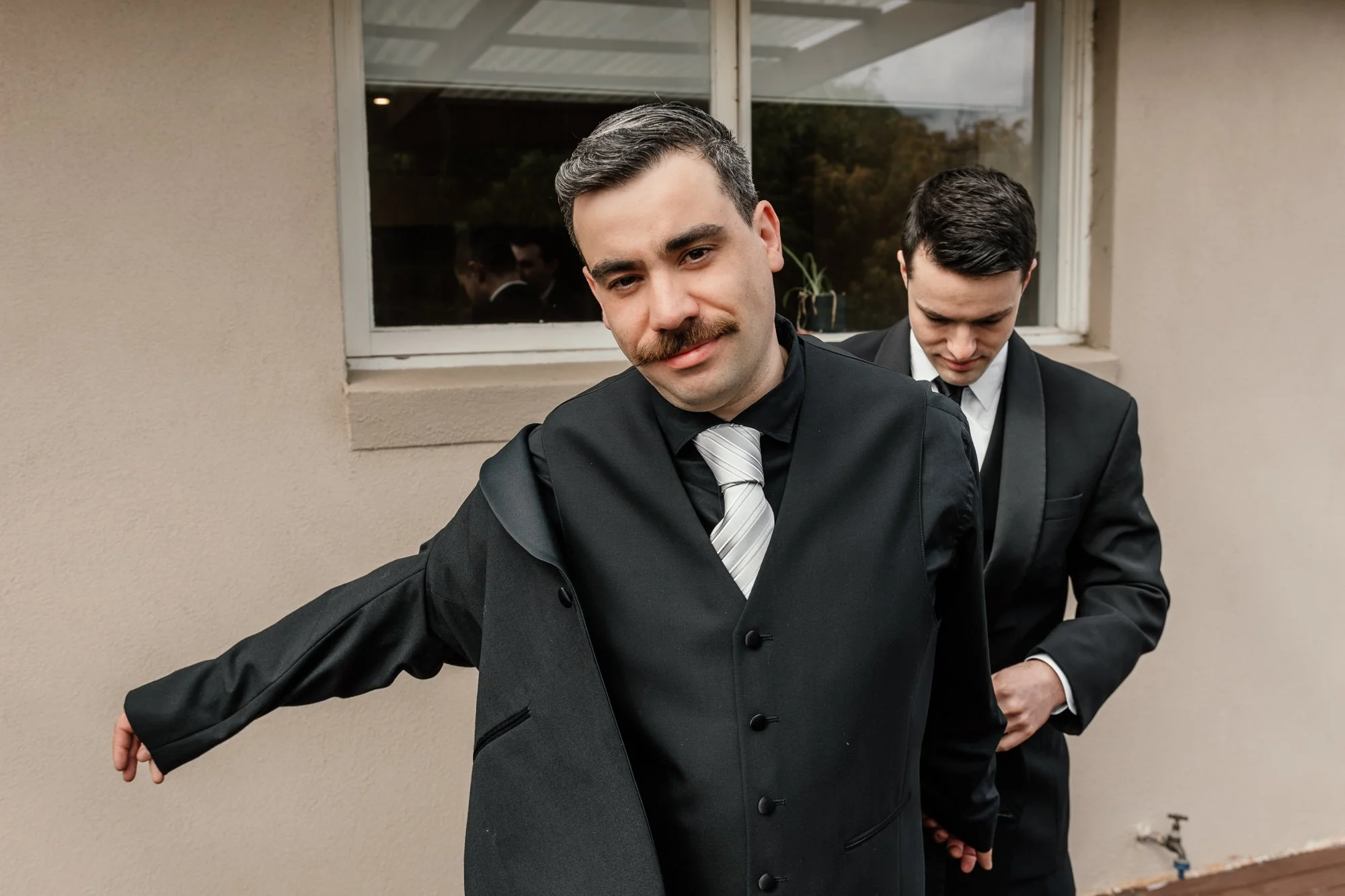 A man with a mustache in a black suit and striped tie standing outdoors against a beige wall with his arm extended, with another man in a black suit behind him, adjusting his sleeve.