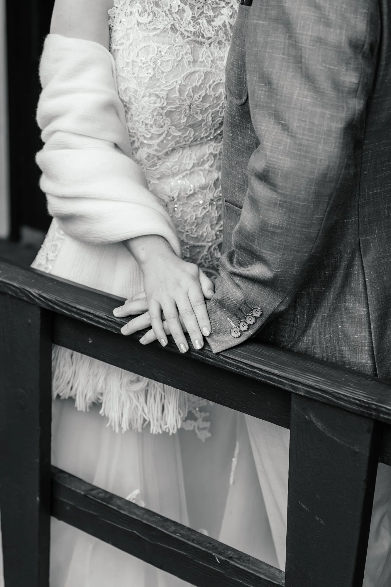 Close-up of a couple holding hands, with a focus on their entwined fingers. The woman wears a lace dress and a cardigan, while the man is in a textured suit jacket. They are standing behind a wooden railing.