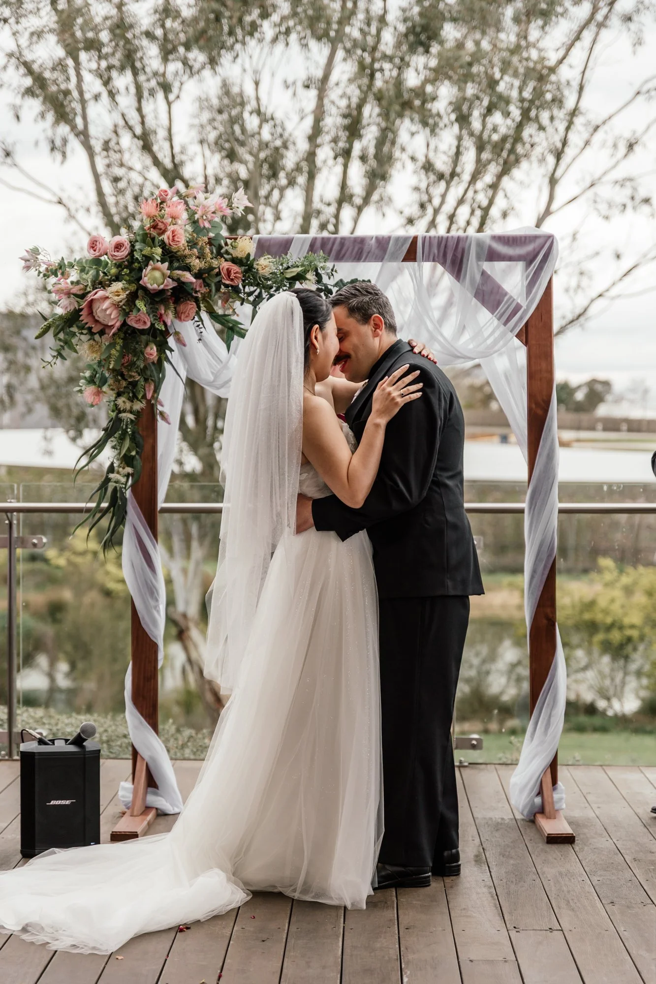 A bride and groom share a kiss under a wedding arch decorated with pink and white flowers and white tulle at an outdoor wedding ceremony. Trees and cloudy sky are in the background.