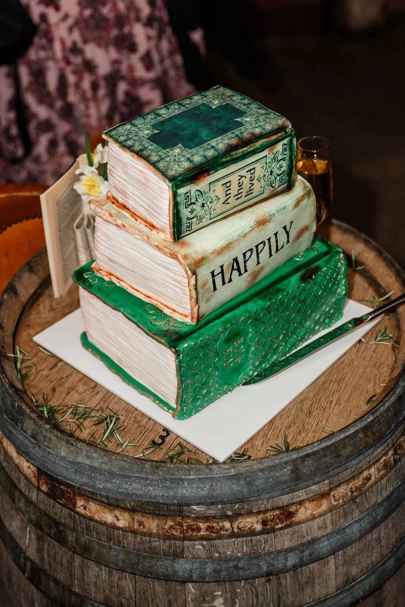 A wedding cake designed to look like a stack of books with titles 'And they lived happily ever after' and 'Happily'. The cake is placed on a wooden barrel with a rustic theme.