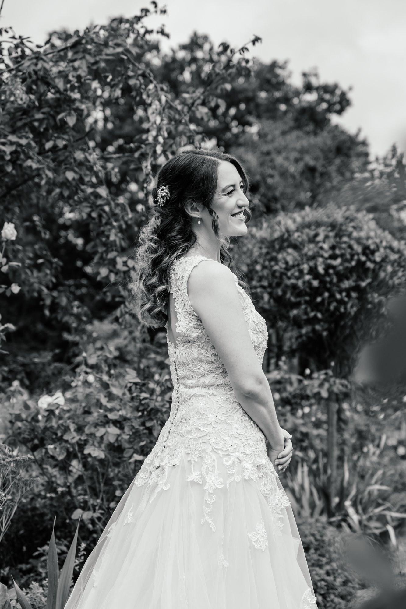 Black and white photo of a smiling woman in a wedding dress outdoors, with trees in the background.