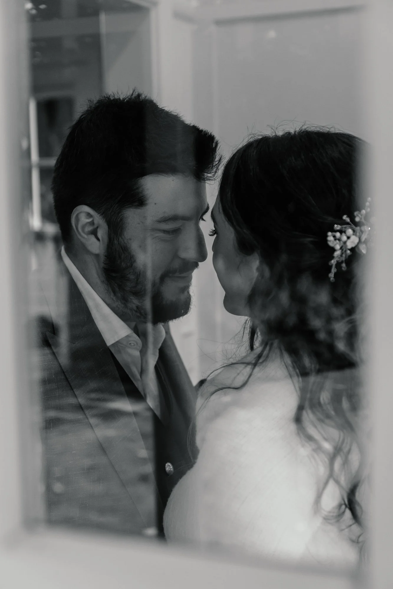 A black and white photo of a couple with foreheads touching, seen through a glass window. The man has dark hair and a beard, wearing a suit, and the woman has long dark hair adorned with a floral hairpiece, wearing a light-colored dress.