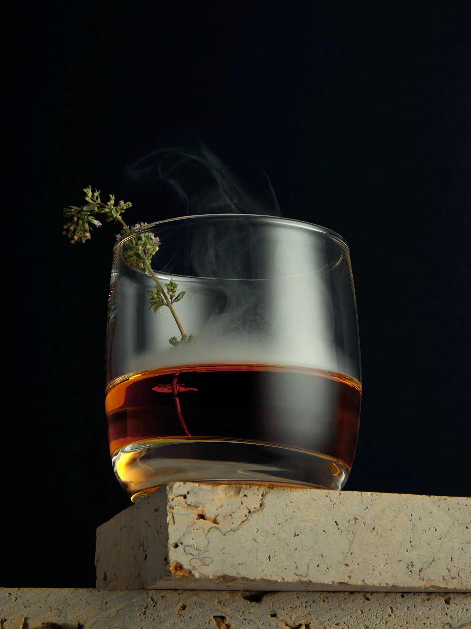 A glass with a warm beverage and a sprig of herbs inside, placed on a rough stone surface against a black background, with steam rising from the drink.