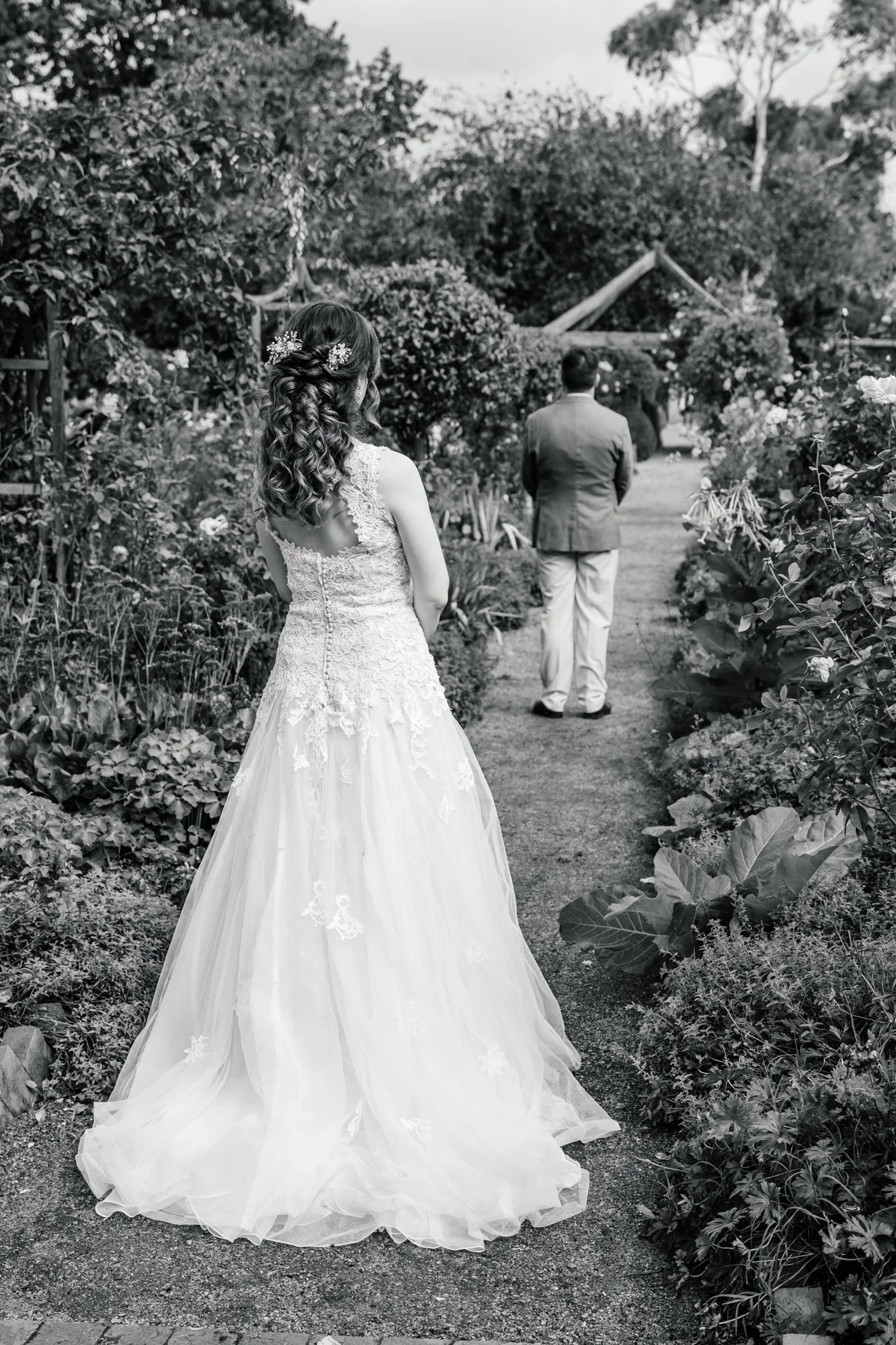 A bride in a wedding dress with curly hair and floral hair accessories stands in a garden, watching a groom in a suit walking away on a garden path, surrounded by greenery and flowers.