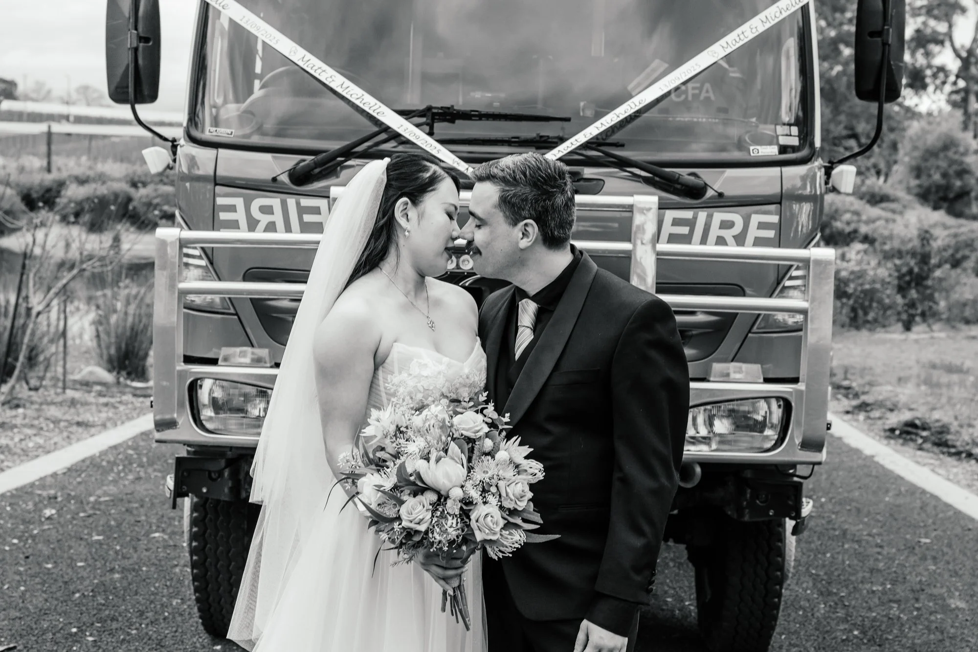 A bride and groom lean their foreheads together in front of a fire truck during a wedding photoshoot. The bride holds a large bouquet of flowers. The scene is outdoors, with trees and an overcast sky in the background.