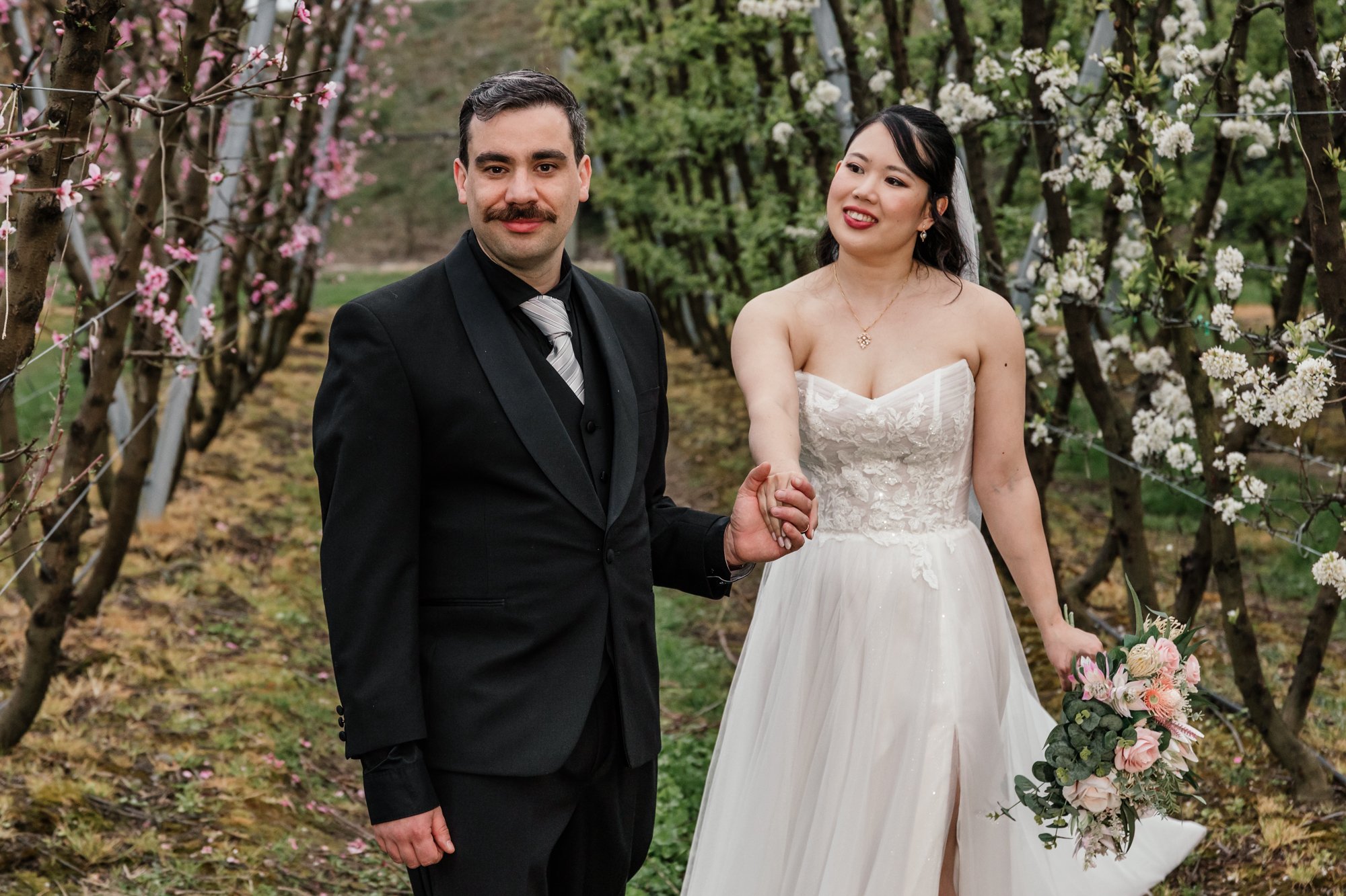 A newlywed couple holding hands in a garden with blooming trees. The groom has dark hair, a mustache, and wears a black suit with a gray tie. The bride has dark hair, is wearing a strapless white wedding dress, and is holding a bouquet of pink and wh
