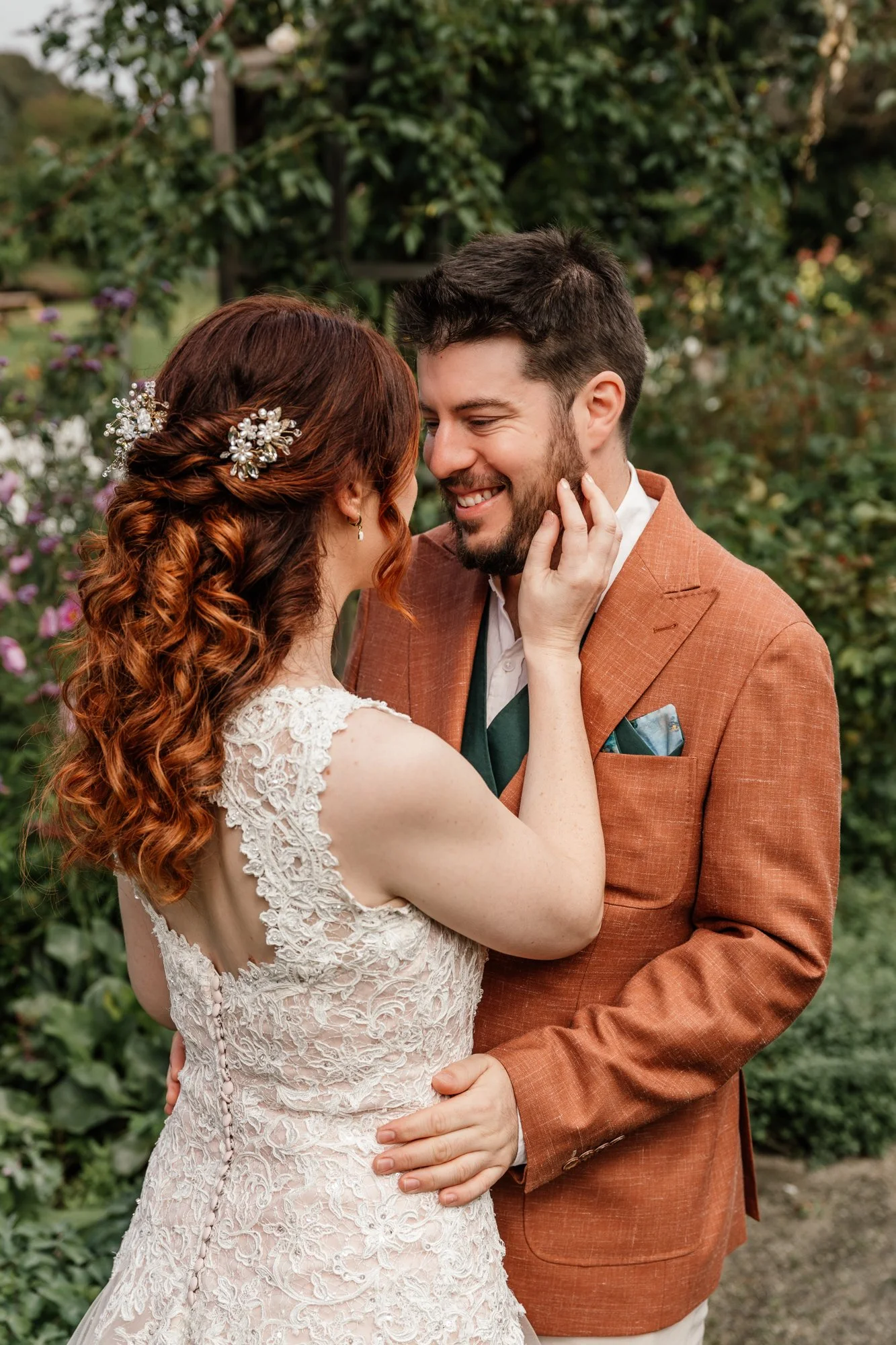 A bride and groom sharing a kiss outdoors. The bride has red curly hair decorated with a pearl and floral hairpiece. The groom has dark hair and a beard, wearing a rust-colored suit with a green tie. They are surrounded by greenery and flowers.