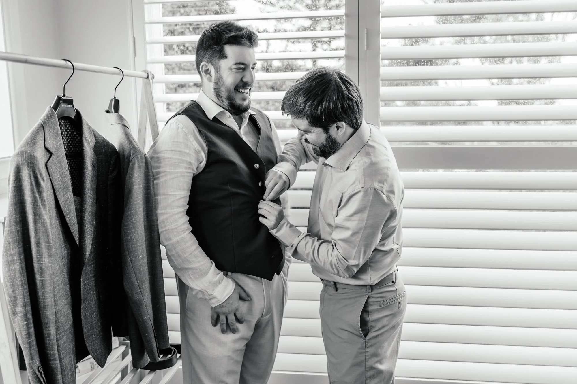 Two men, one helping the other with a vest in a clothing store, with suits hanging on a rack to the side and window blinds in the background.