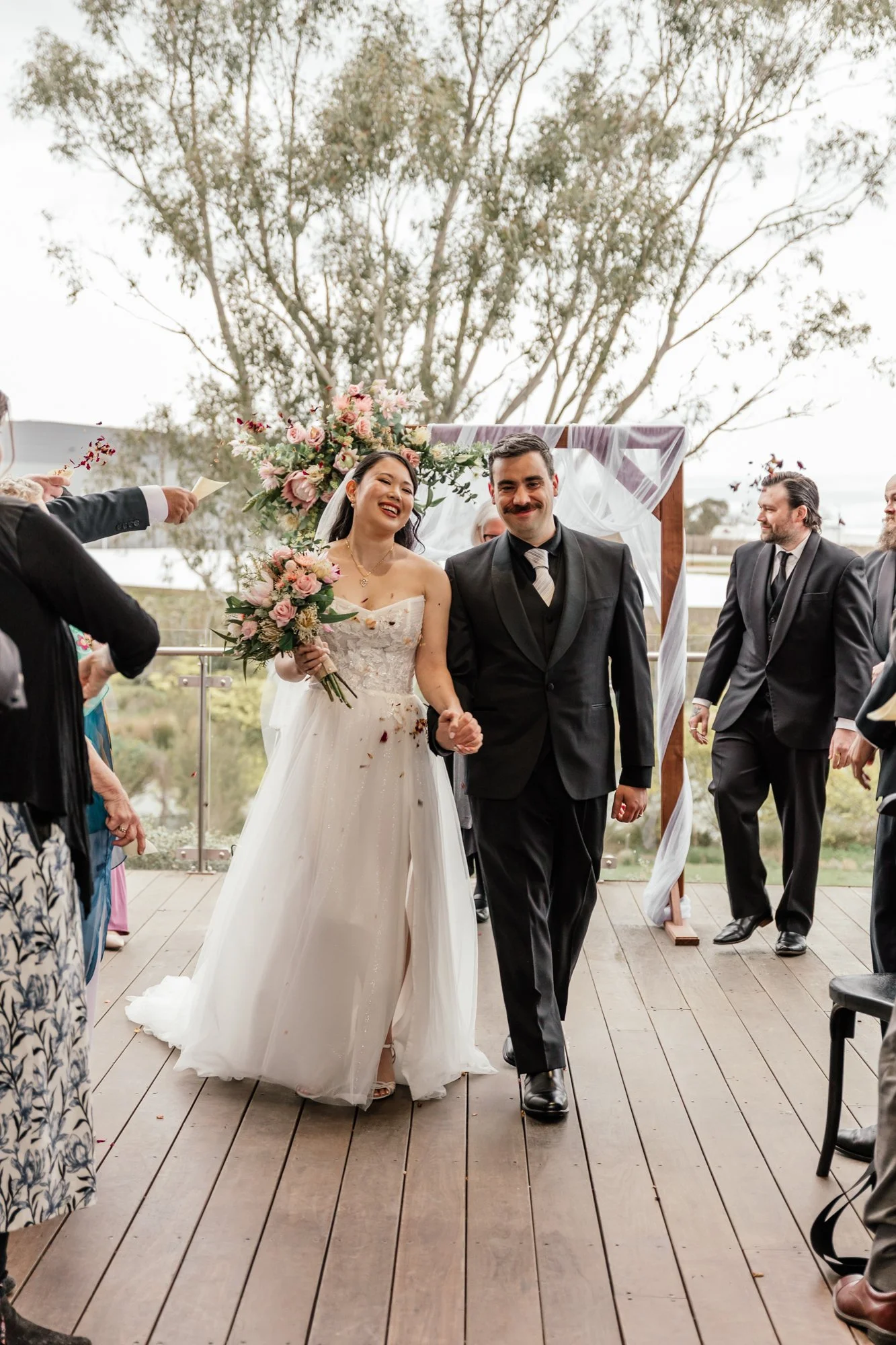 A bride and groom smiling and holding hands during their wedding ceremony outdoors, with friends and family around them.