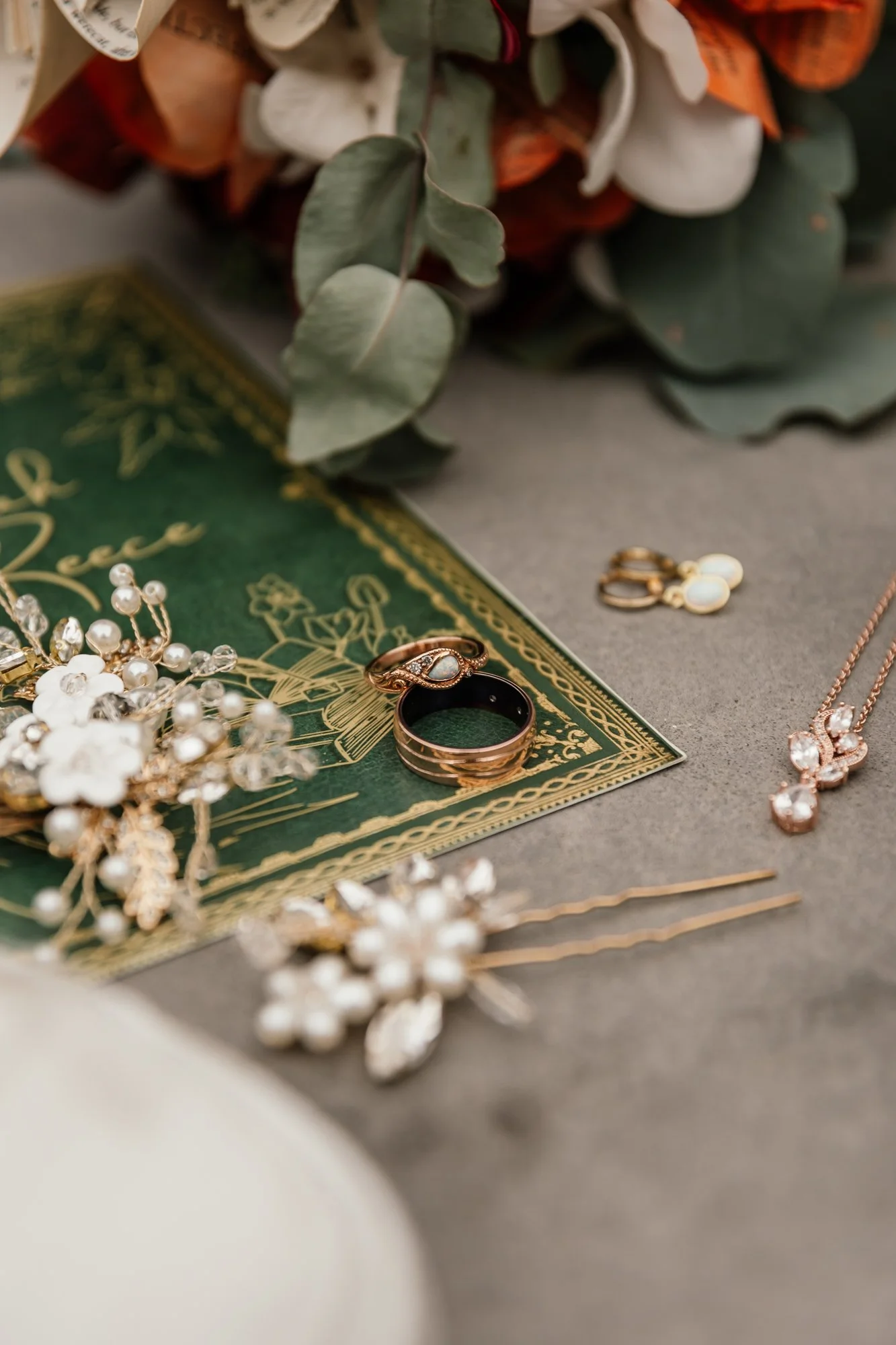 Close-up of jewelry including a pearl and gold necklace, a gold ring with an opal, a pair of drop earrings with opals, and a gold necklace, arranged on a green decorative card, with a bouquet of white and orange flowers and eucalyptus leaves in the b
