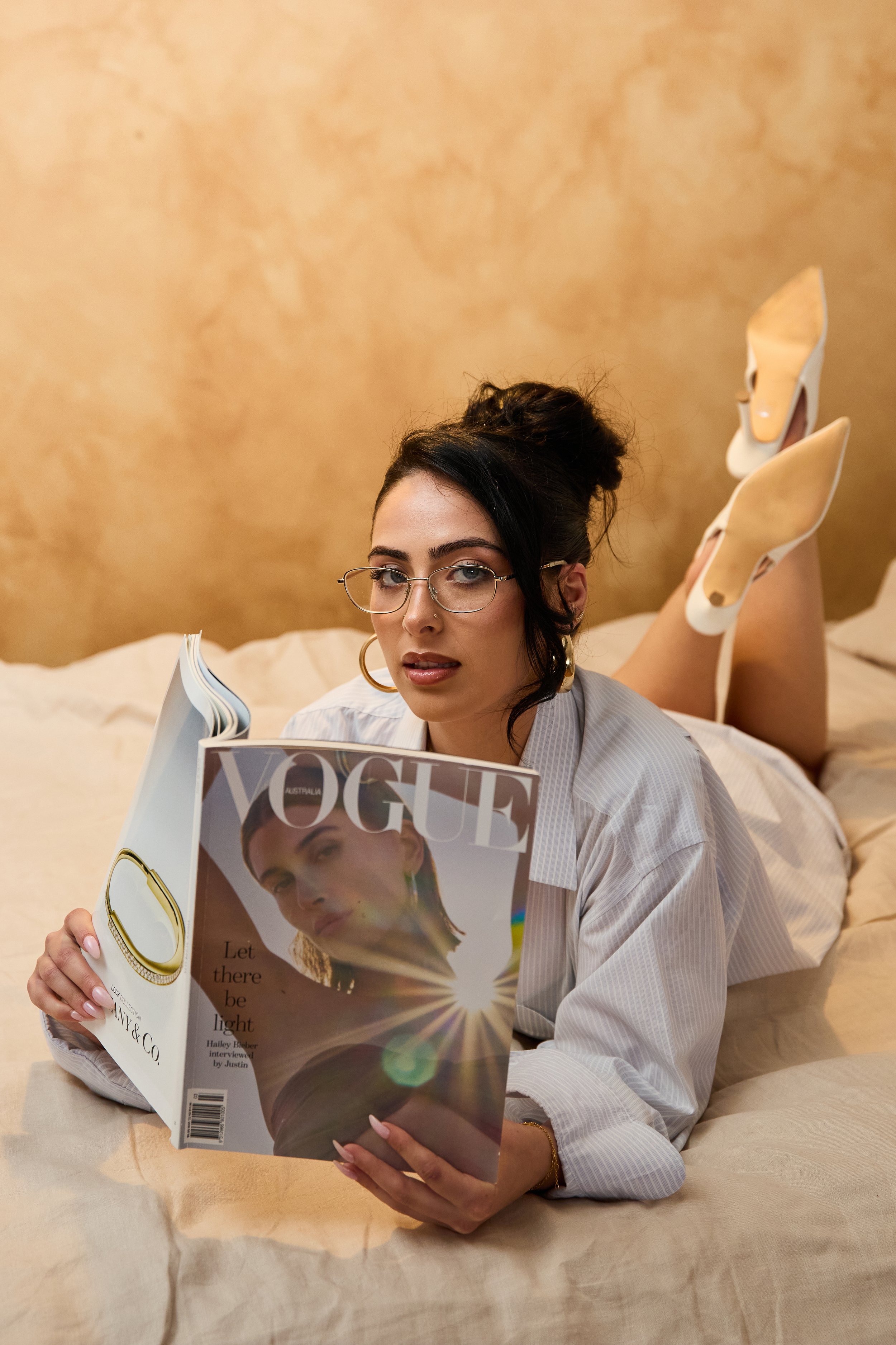 Woman with dark hair, glasses, and gold hoop earrings lying on a bed, holding a Vogue magazine featuring a woman with short hair and multiple earrings on the cover.