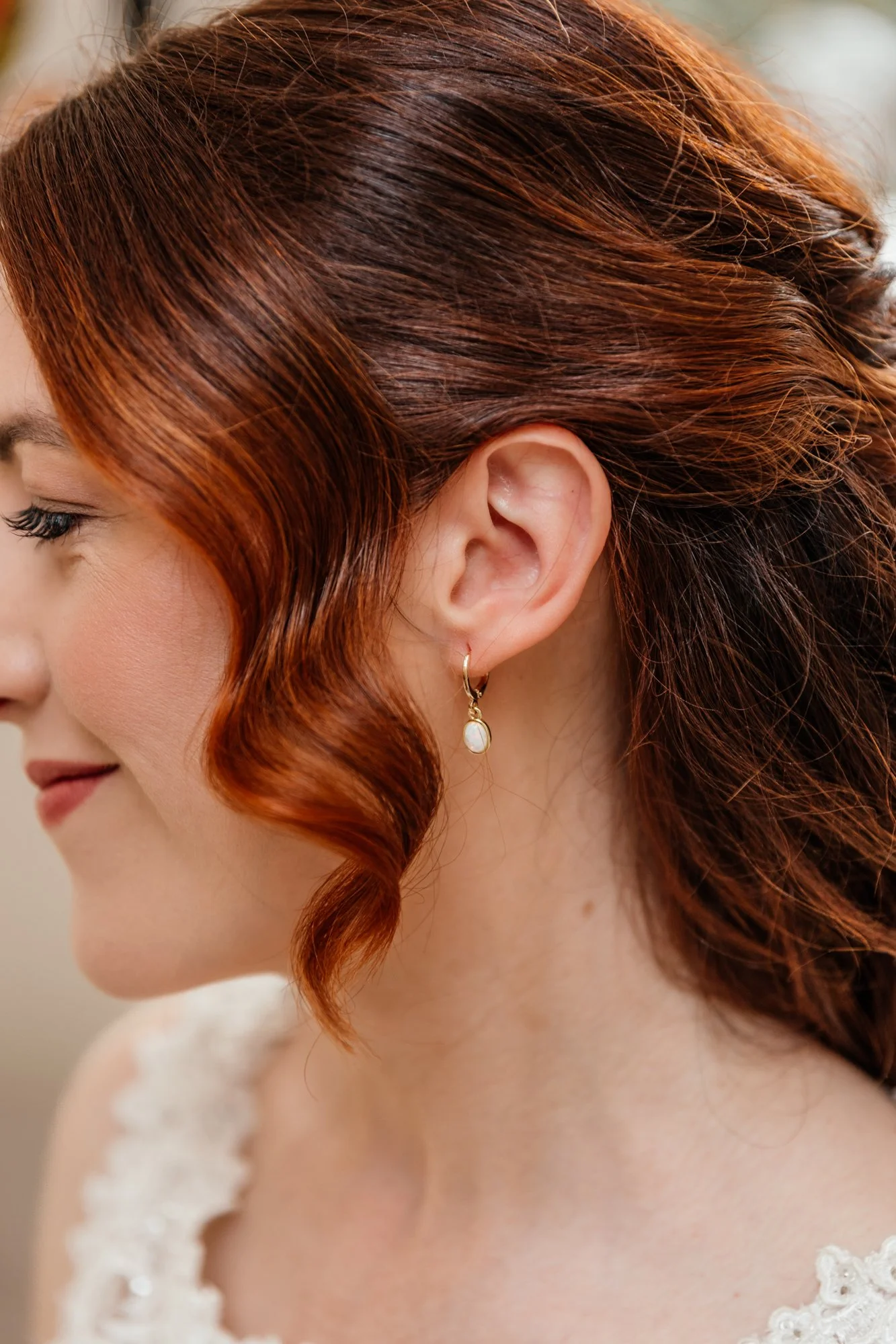 Close-up of a woman with red, wavy hair wearing a white lace dress and a dangling pearl earring.