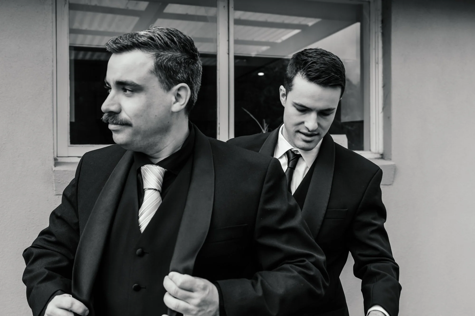 Two men wearing suits and ties, standing outside near a building with a window, engaged in conversation, in black and white image.