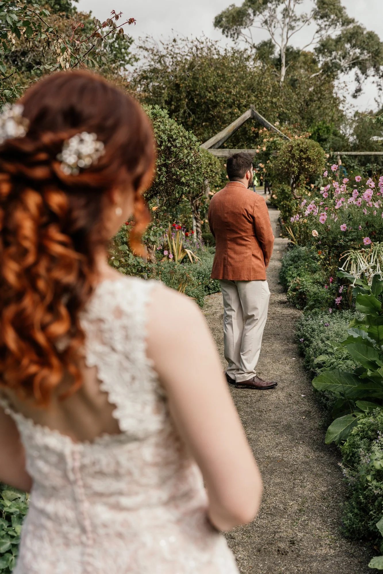 A woman with curly red hair decorated with white floral accessories is in the foreground, slightly out of focus. In the background, a man with dark hair and a beard, wearing a brown blazer and beige pants, stands on a garden path surrounded by bloomi