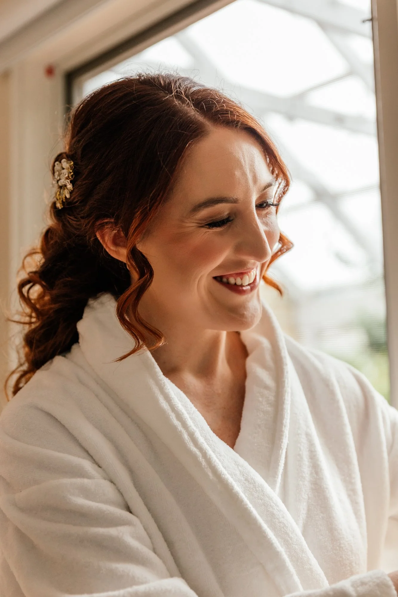 A woman with red hair styled in loose curls, wearing a white bathrobe, smiling softly while looking downward at an indoor window.