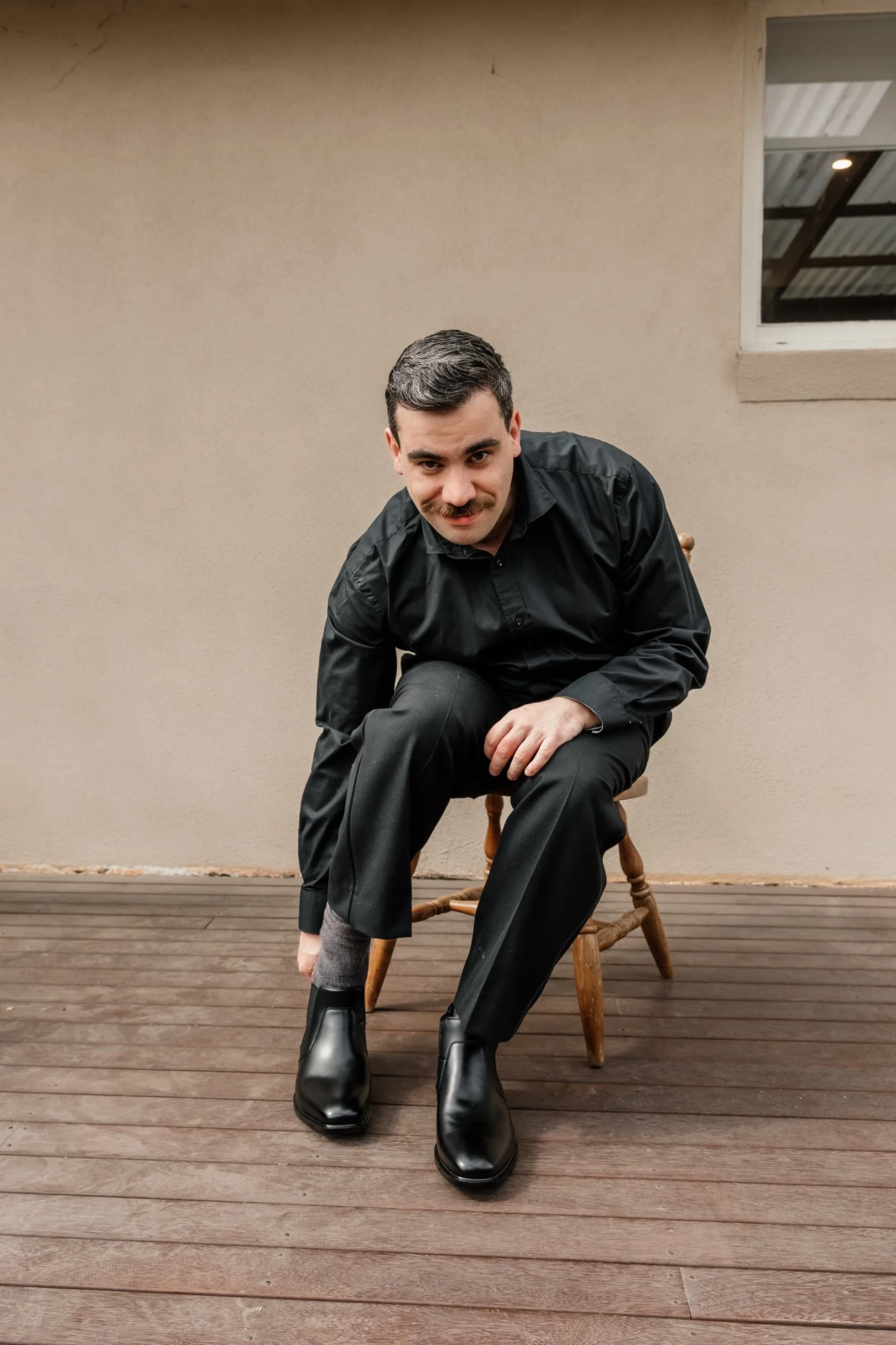 A man with dark hair, a mustache, and light skin is sitting on a wooden chair on a wooden deck, adjusting his shoe. He is wearing a black dress shirt, black pants, and black boots. The background includes a beige wall with a window and a ceiling with