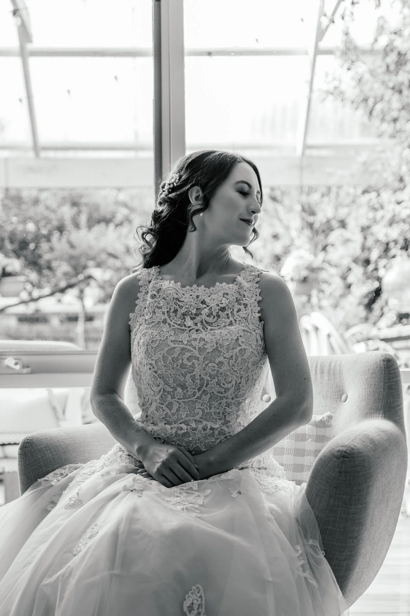 Black and white photo of a woman in a wedding dress sitting on a couch, with a large window and trees in the background.