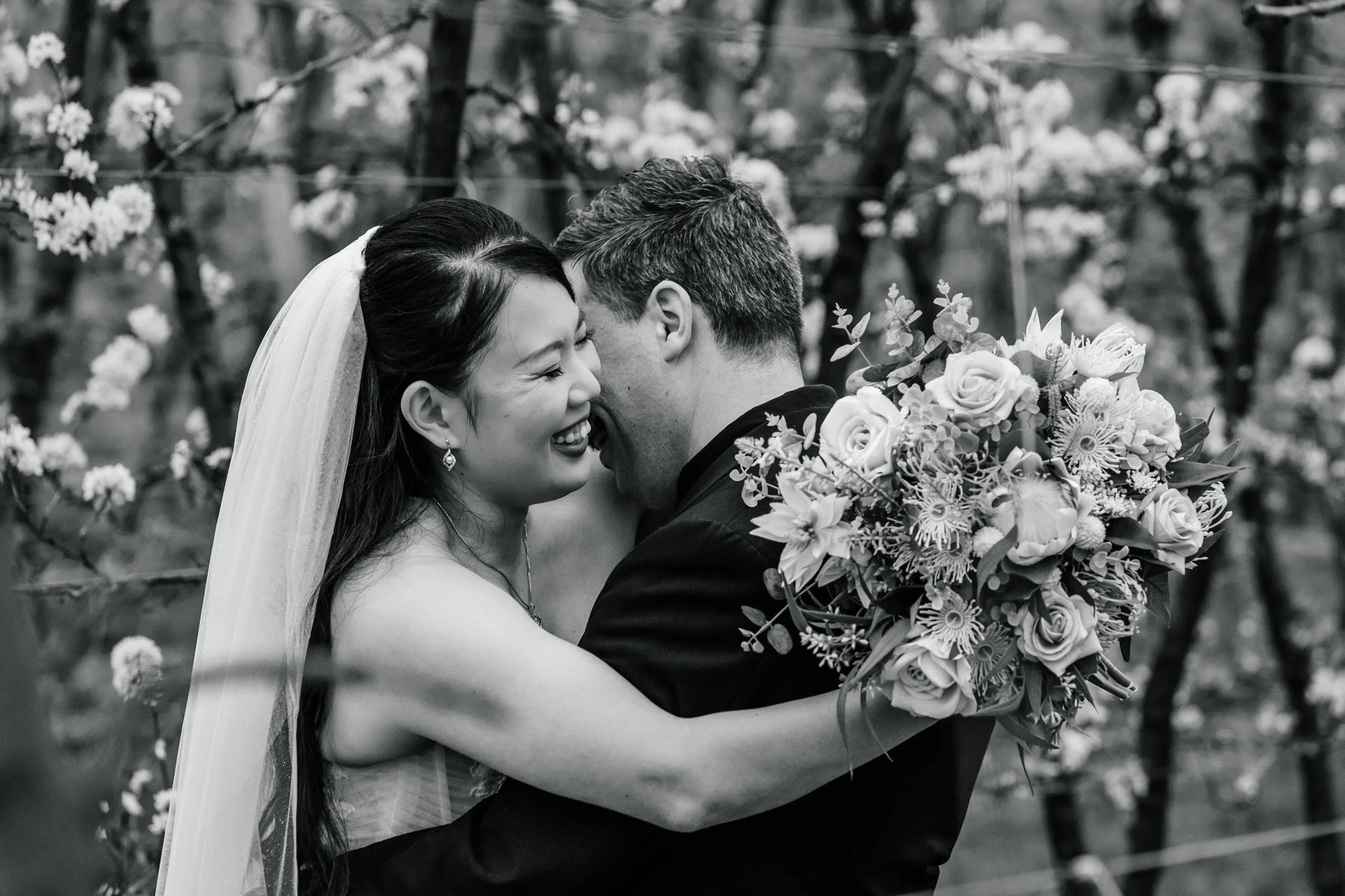 A smiling bride and groom embrace outdoors during a wedding, with the groom holding a large bouquet of flowers. They are surrounded by blooming trees.