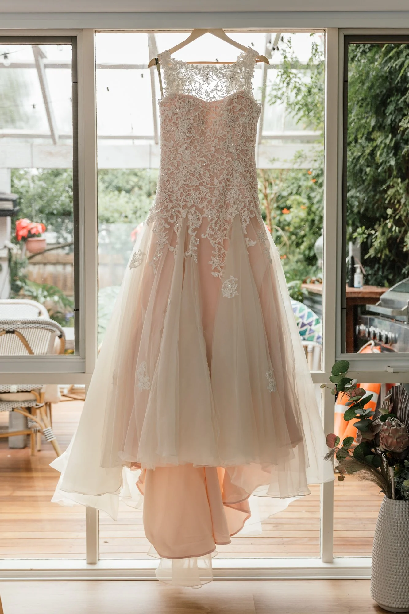 A wedding dress hanging in front of a large window with greenery outside. The dress has lace details on the bodice and a flowing skirt.