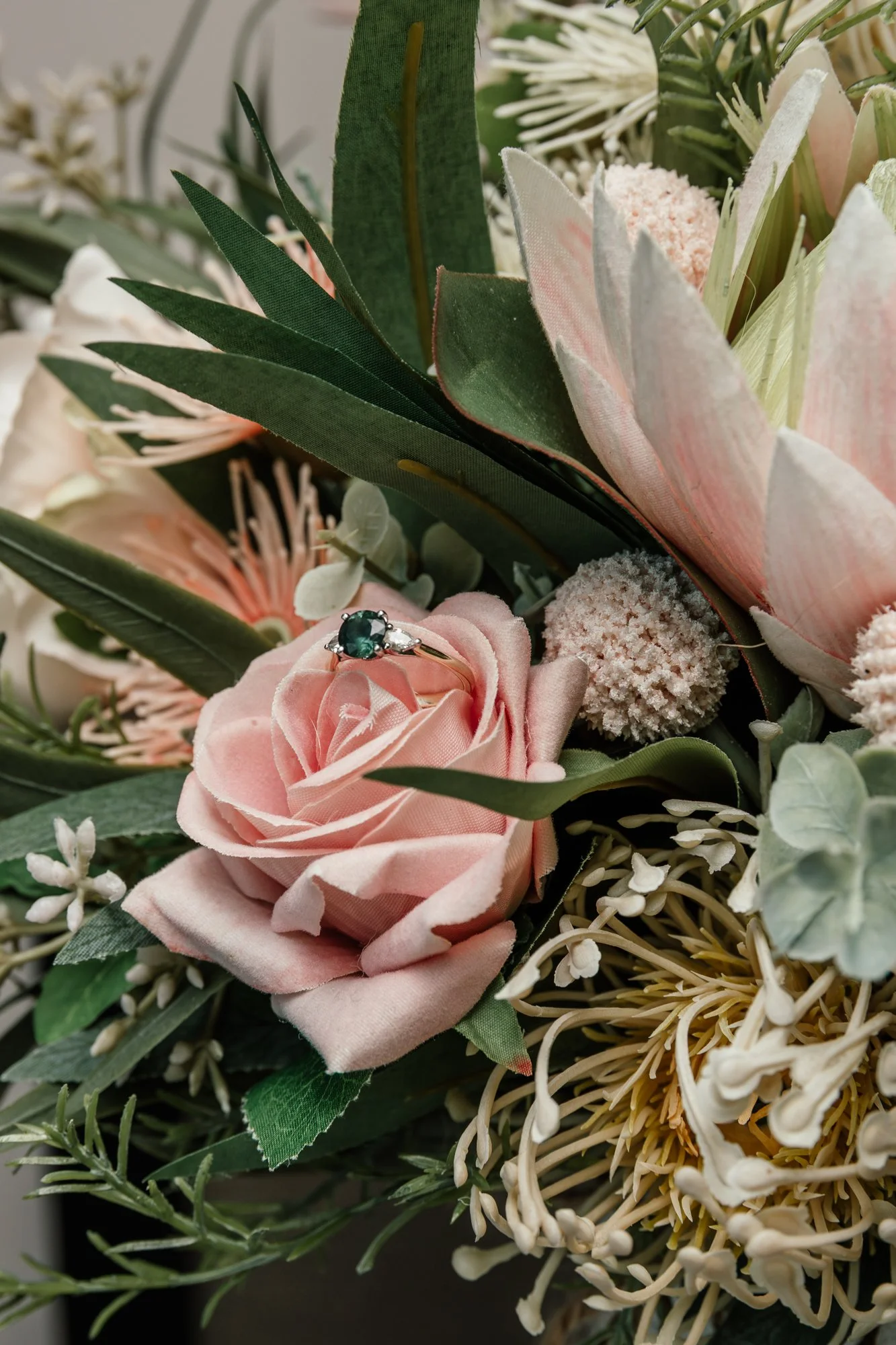 A bouquet of flowers featuring a pink rose, various greenery, and white flowers with a small ring with a green gemstone resting on the pink rose.