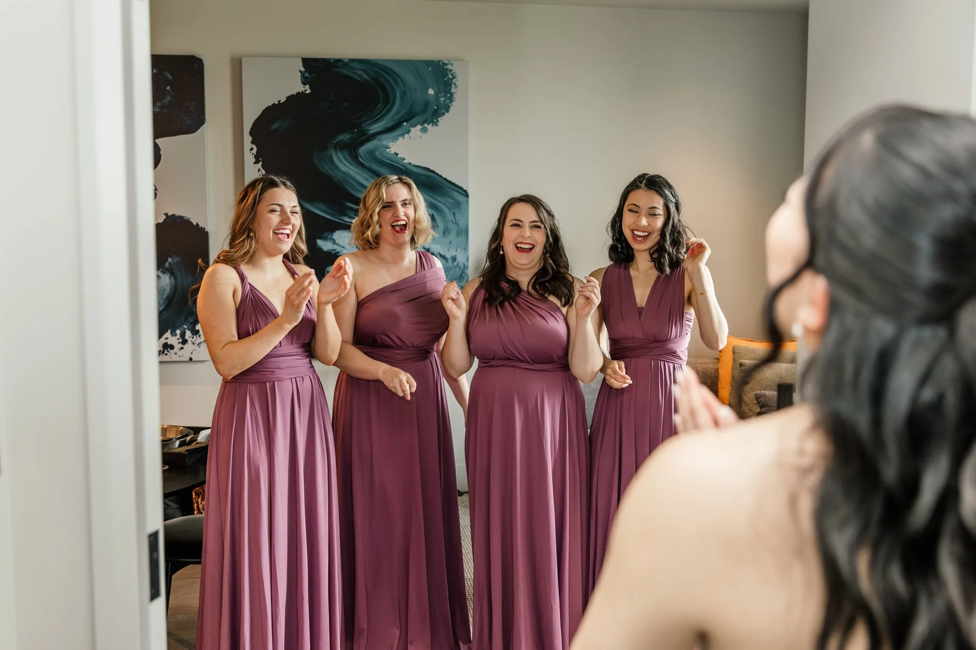 Four women in matching mauve bridesmaid dresses smiling and laughing while standing together indoors, with artwork behind them and a woman in the foreground with dark hair and glasses