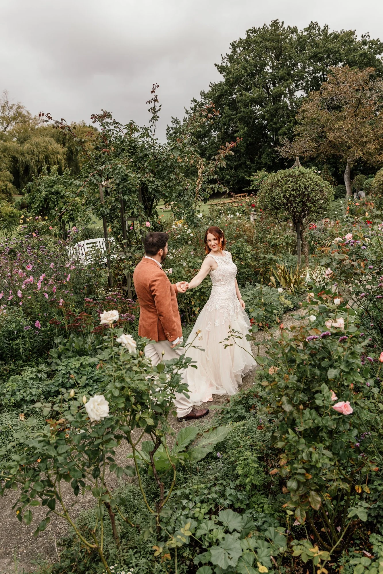 A bride and groom holding hands in a lush garden filled with blooming roses and greenery, overcast sky in the background.