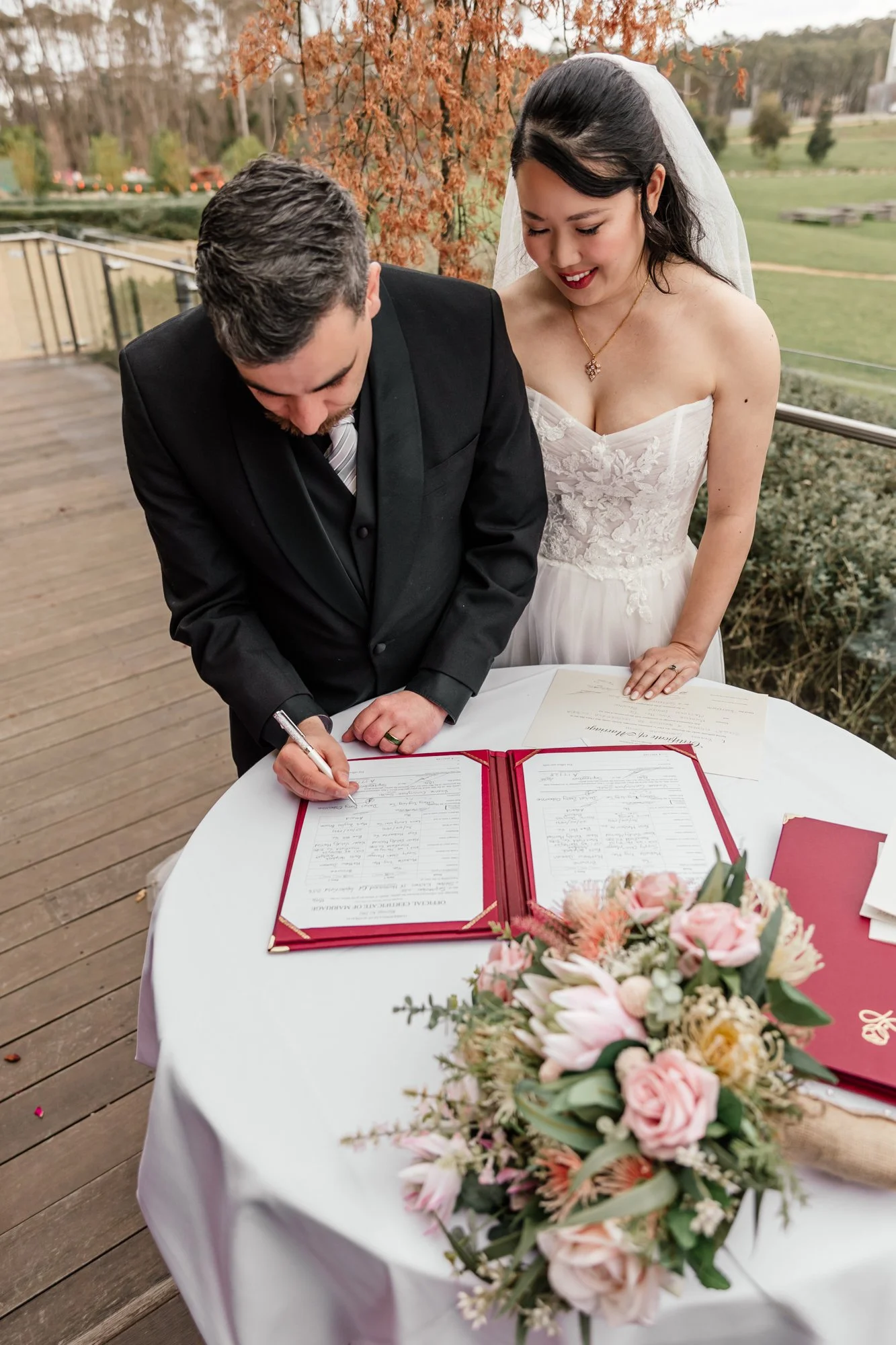 A bride and groom signing a marriage certificate at an outdoor wedding ceremony, with a bouquet of pink and white flowers on a table in the foreground and trees in the background.
