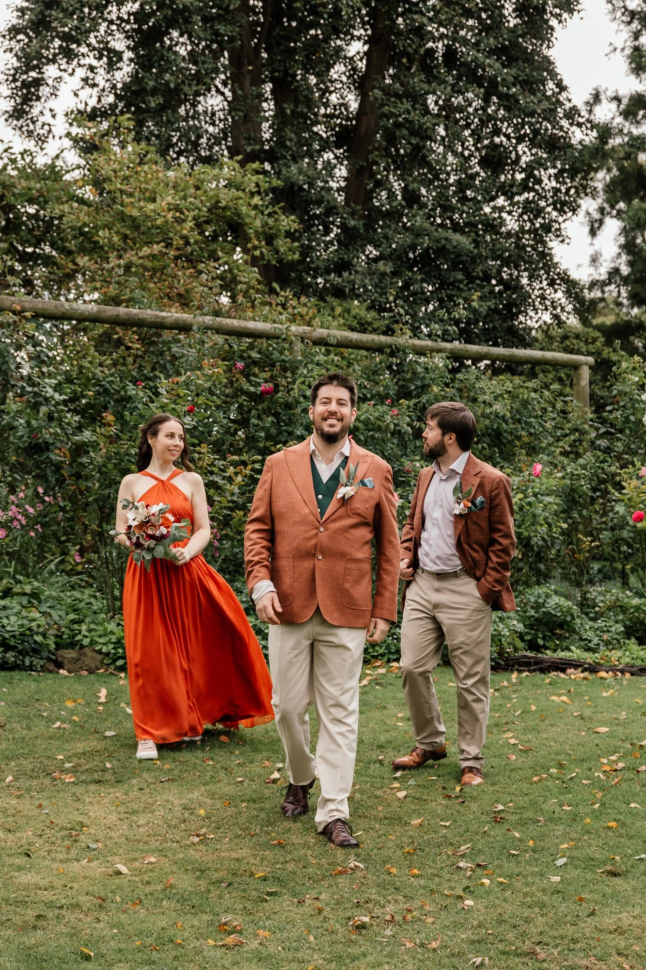 A group of three people walking on a grassy area outdoors. The man in the front is smiling and wearing a brown jacket with beige pants. The woman on the left is holding a bouquet of flowers, wearing a flowing red dress. The man on the right is wearin