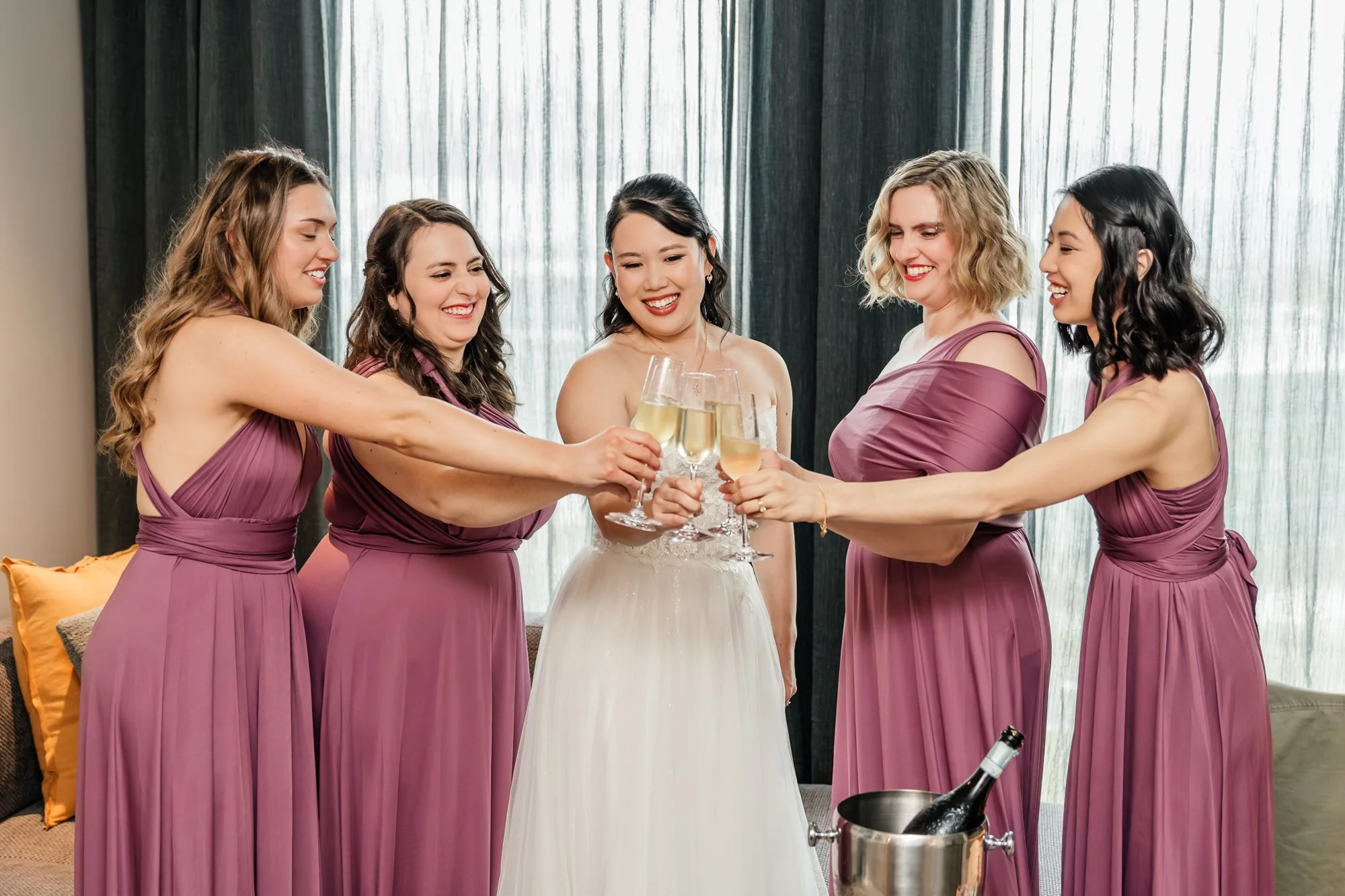A group of five women in matching purple dresses holding champagne glasses in a toast, with a bride in a white gown in the center celebrating at a gathering or wedding reception.