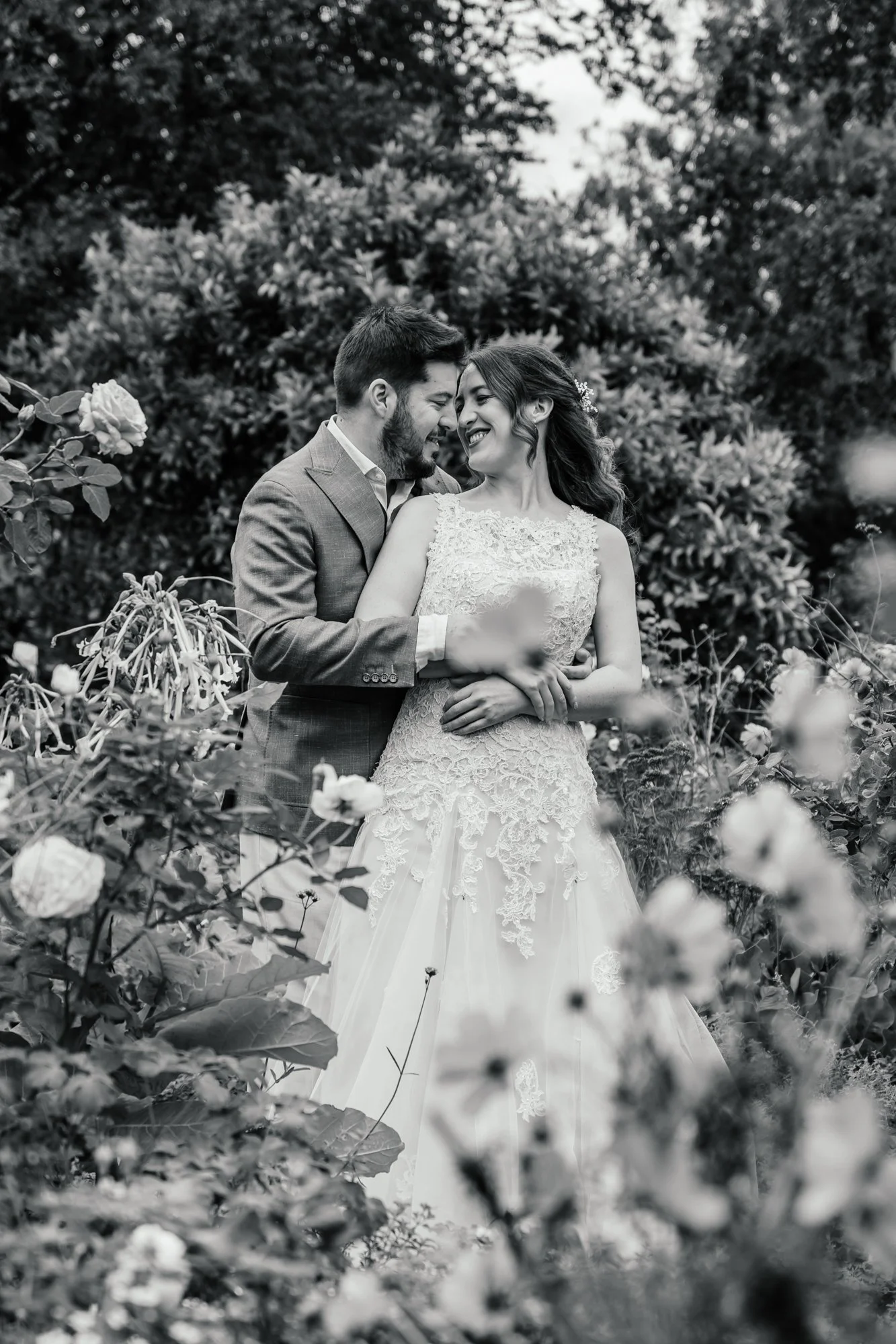 A black and white photo of a newlywed couple smiling and touching foreheads in a garden with blooming flowers and bushes.