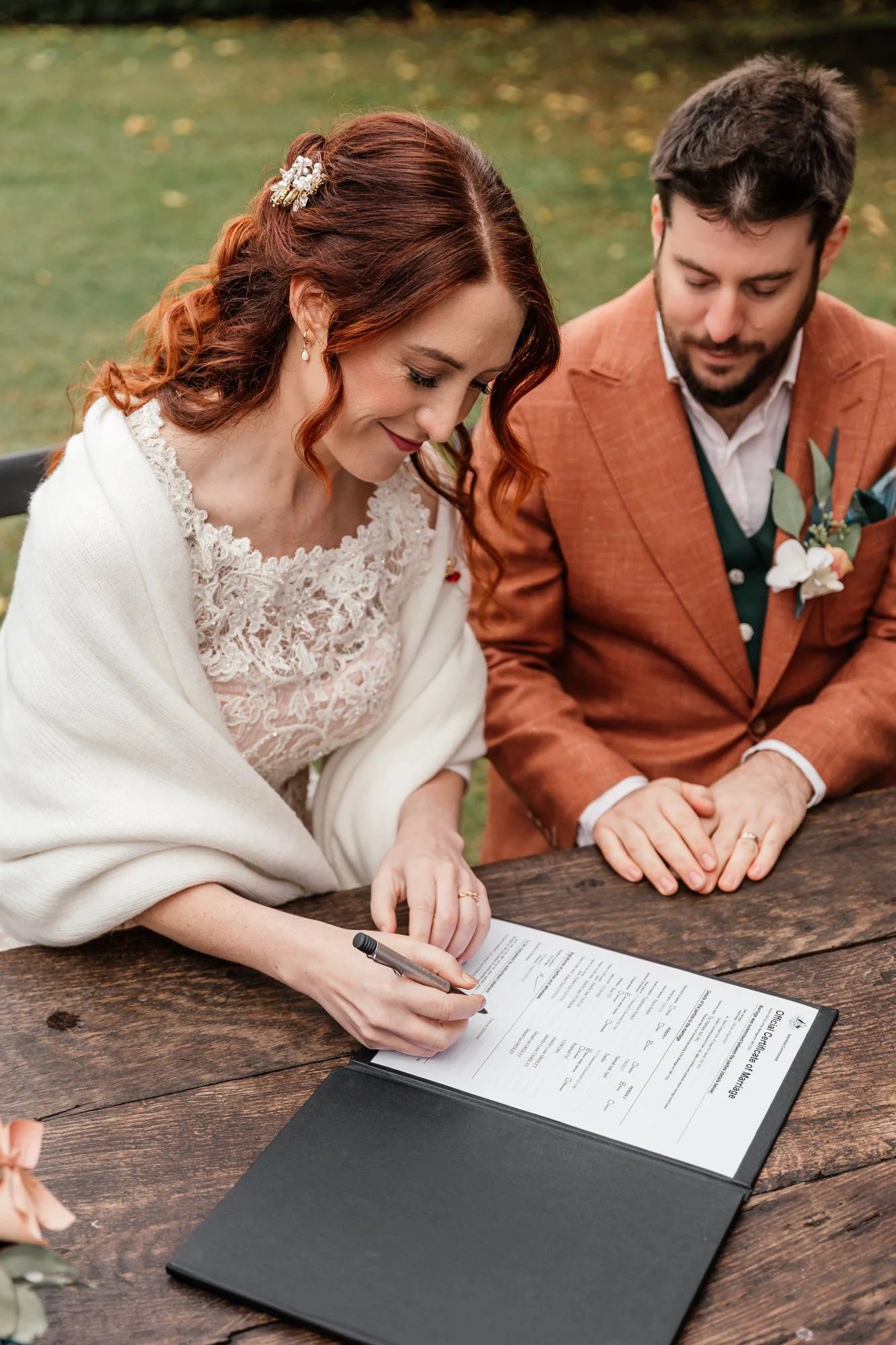 A woman with red hair, wearing a white lace dress and a hair accessory, signs a document at a wooden table, with a man in a brown suit observing.