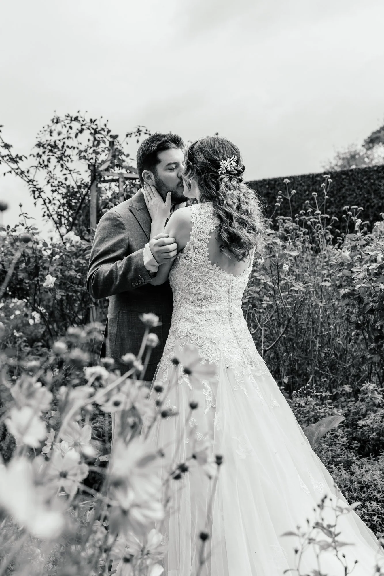 A black-and-white photo of a bride and groom sharing a kiss in a garden with flowers and a hedge in the background.