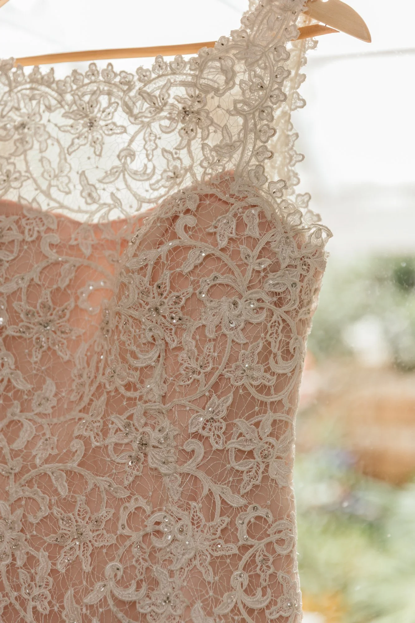 A close-up of a lace dress with intricate embroidery and floral patterns, hanging on a wooden hanger, with a blurred outdoor view in the background.