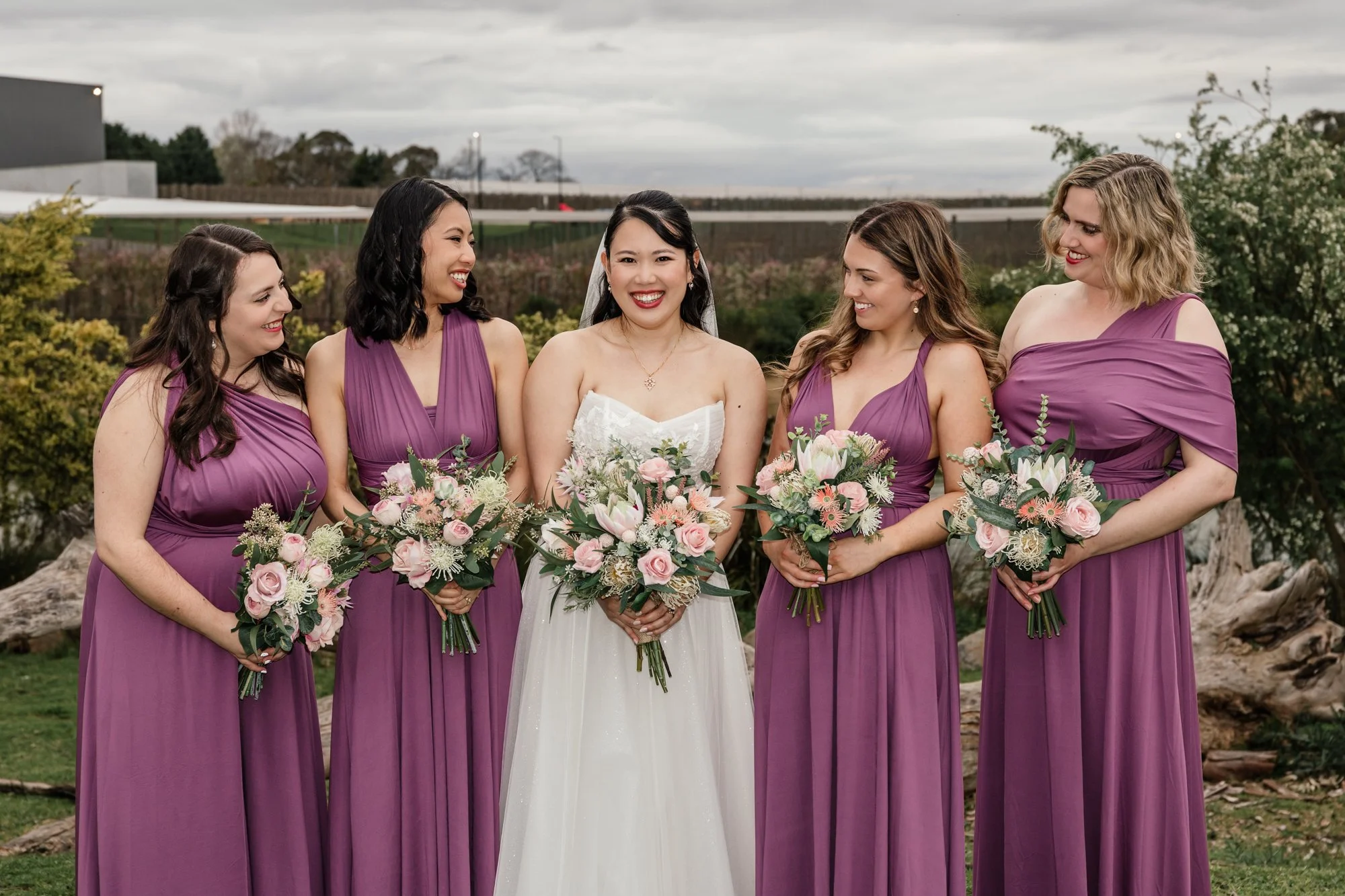 Group of five women, dressed in purple bridesmaid dresses except for the bride in a white wedding gown, standing outdoors with bouquets, overcast sky in the background.