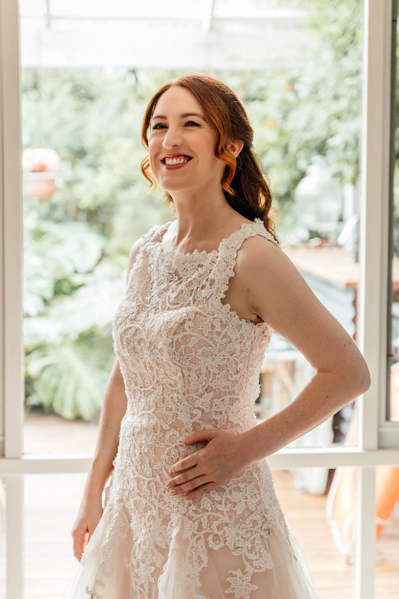 A woman with red hair smiling in a wedding dress indoors near a window with a garden view.