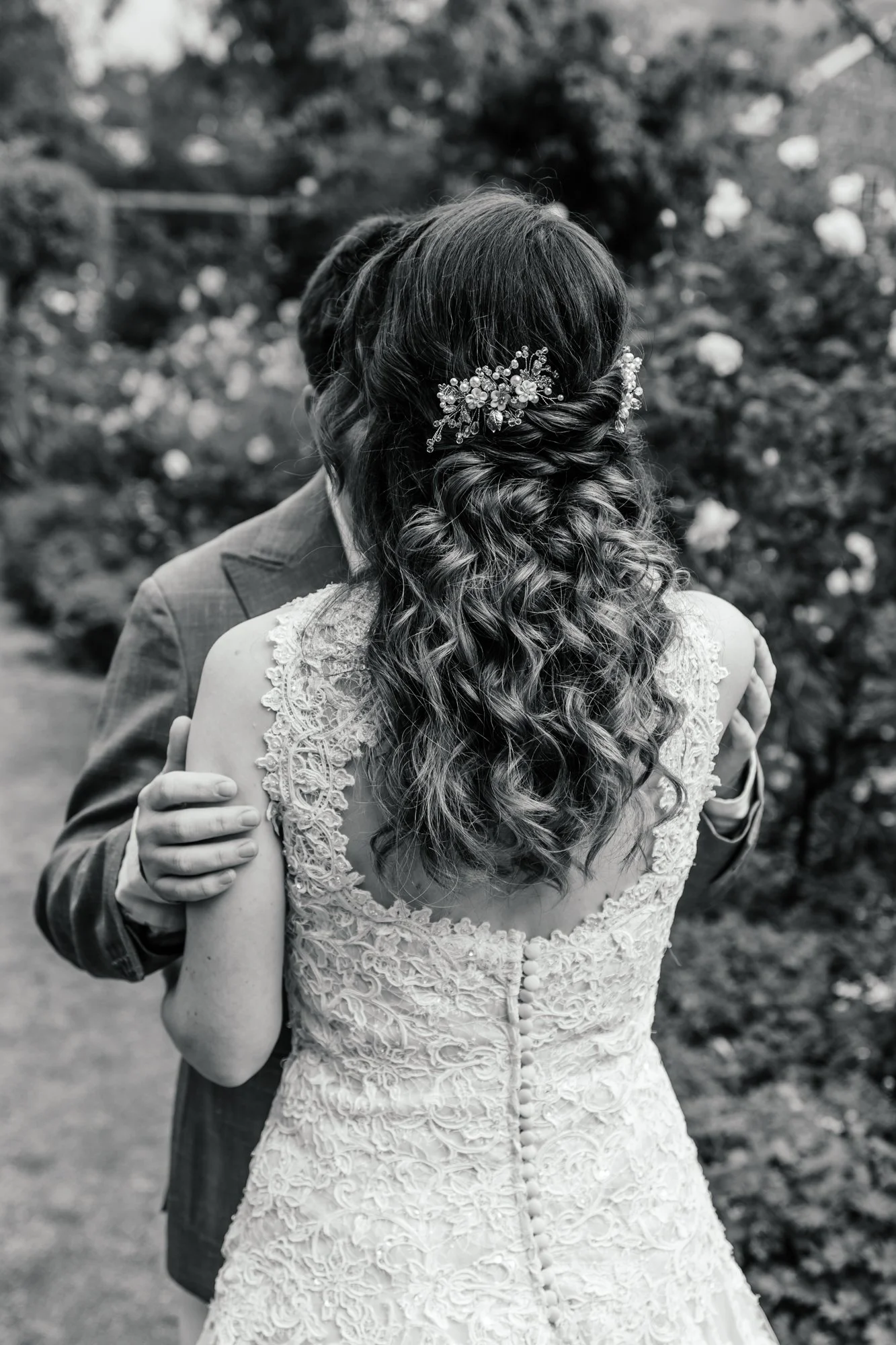 Black and white photo of a bride and groom embracing each other outdoors, focusing on the bride's intricately detailed lace wedding dress and her wavy hairstyle adorned with a floral hair accessory.