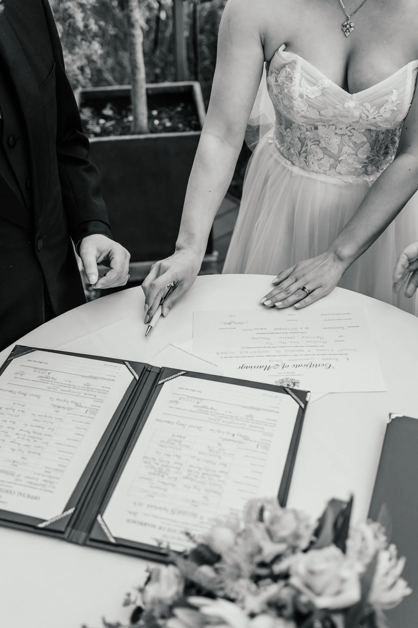A wedding ceremony signing table with documents and a pen, a bride in a lace dress, a groom in a suit, and floral decorations.