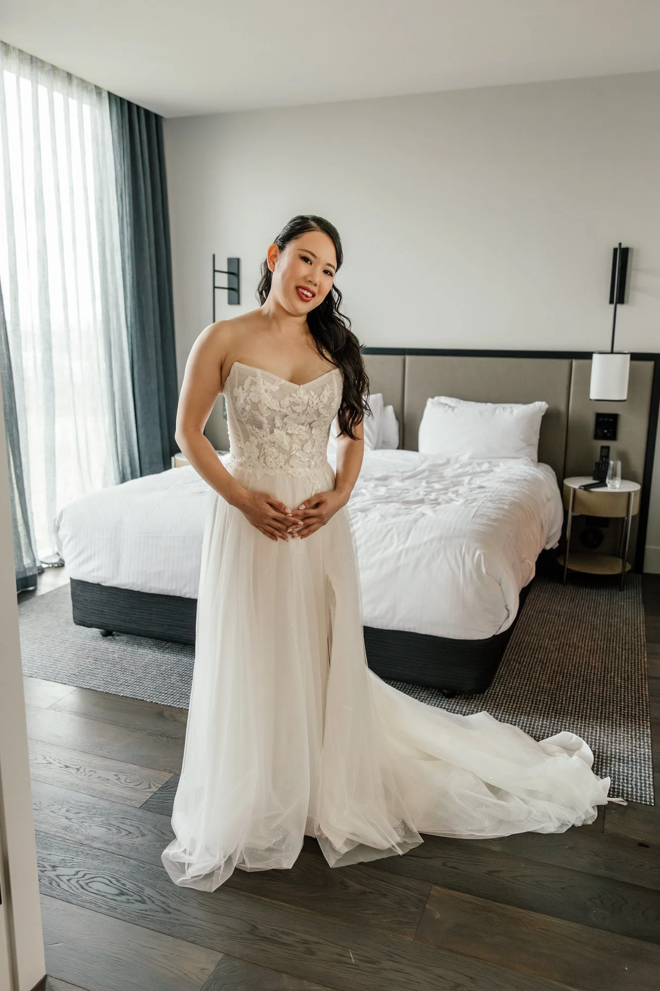 A woman in a strapless white wedding dress standing in a hotel room, smiling and holding her stomach, with a bed and large window with curtains in the background.