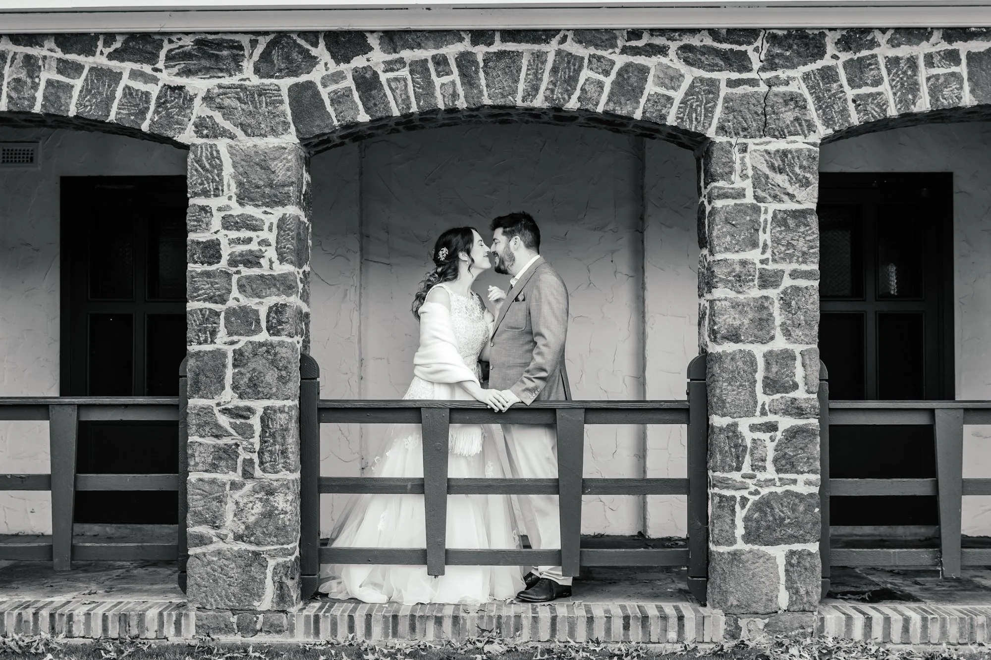 Black and white photograph of a bride and groom standing close together on a small porch, facing each other with noses touching. The bride wears a lace wedding dress with a floral hair accessory, and the groom wears a suit. They are framed by brick a