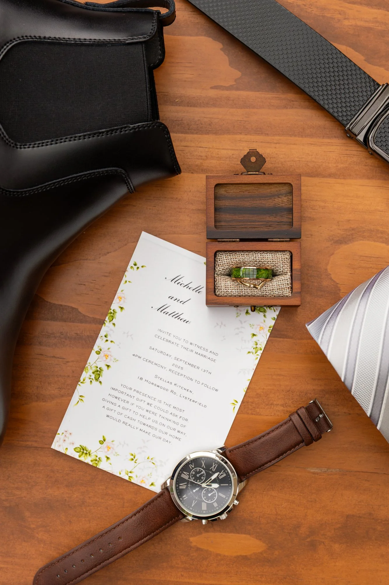 A wedding invitation card with floral design and text about the wedding of Michelle and Matthew, placed on a wooden surface. Surrounding items include a black bag, a gray textured belt, a wooden jewelry box with a ring, a wristwatch with a brown leat
