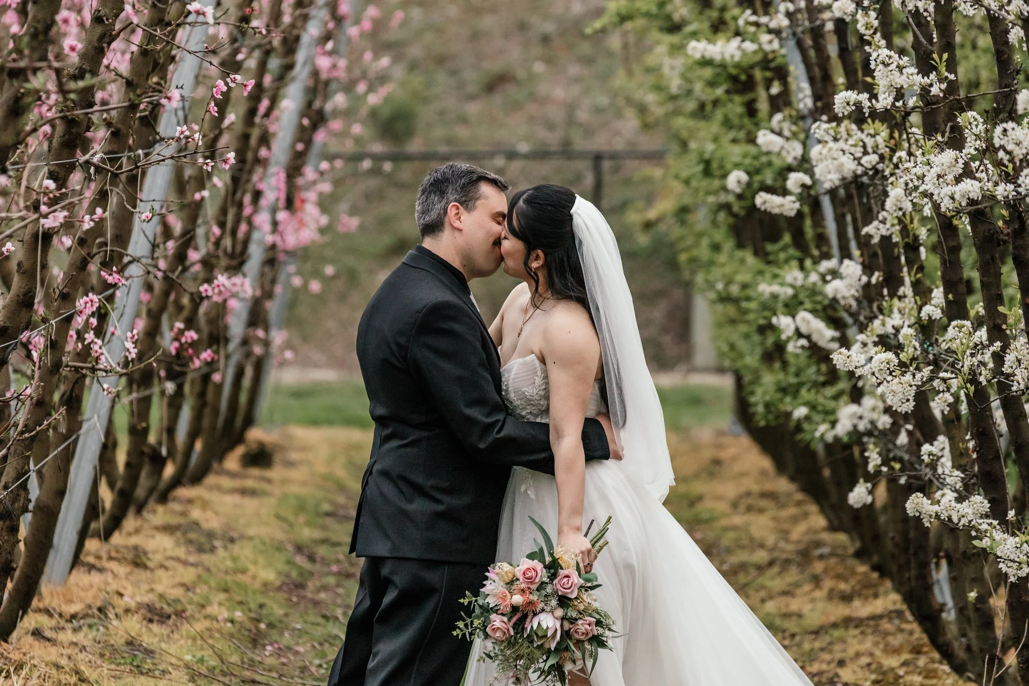 A bride and groom share a kiss in a garden aisle with blossoming trees, the bride holding a bouquet of pink and white flowers and the groom in a black suit.