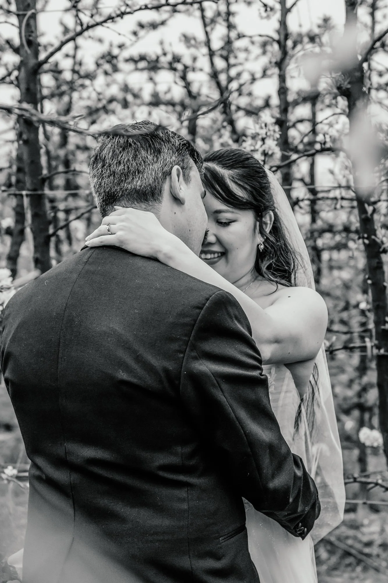 A black and white photograph of a bride and groom embracing, smiling, and closing their eyes during a wedding ceremony in an outdoor setting with trees and branches in the background.