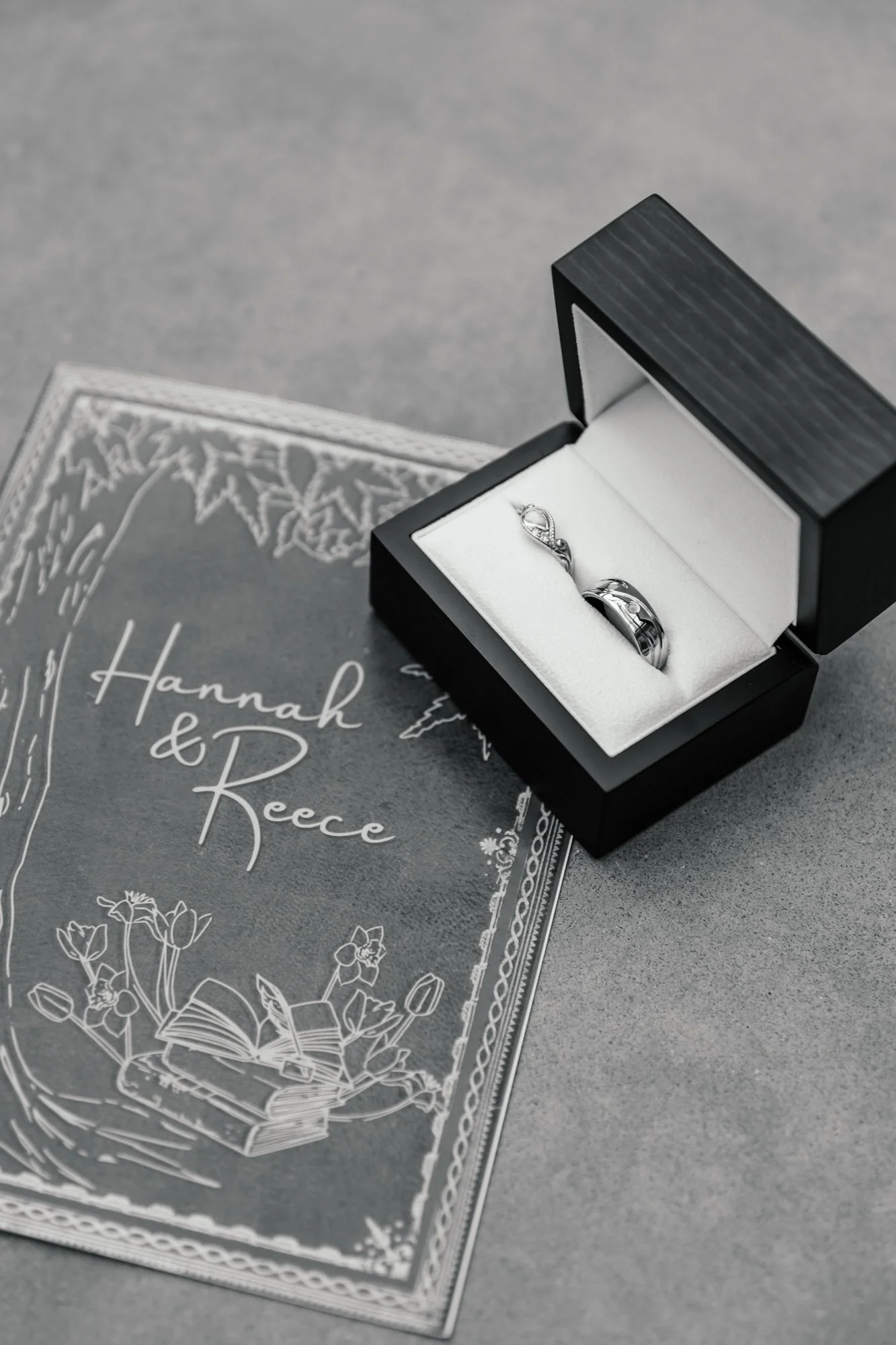 A black jewelry box with rings inside, placed on top of a printed wedding invitation card with floral designs.